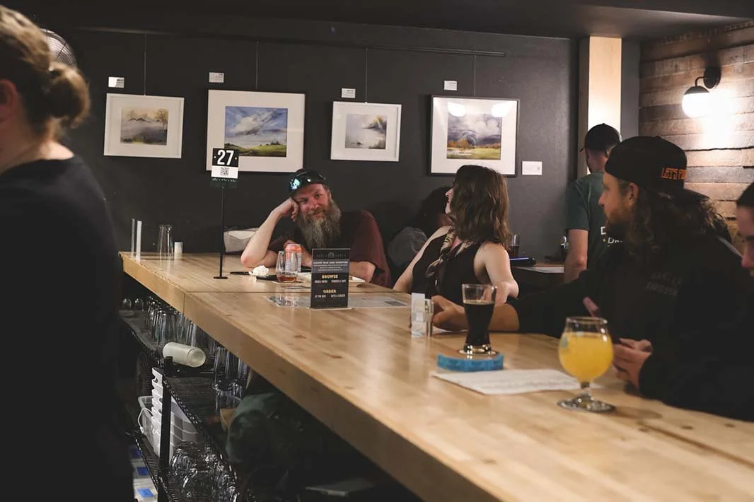 People sitting at a bar with drinks, talking and smiling, with paintings on the wall behind them.