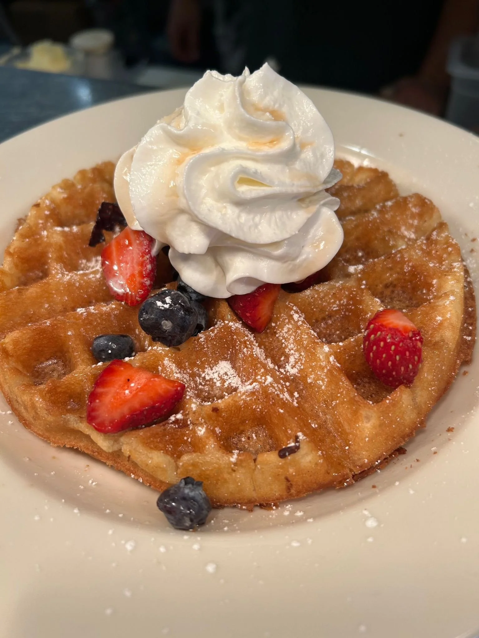 Close-up of a waffle topped with whipped cream and fresh mixed berries, sprinkled with powdered sugar.