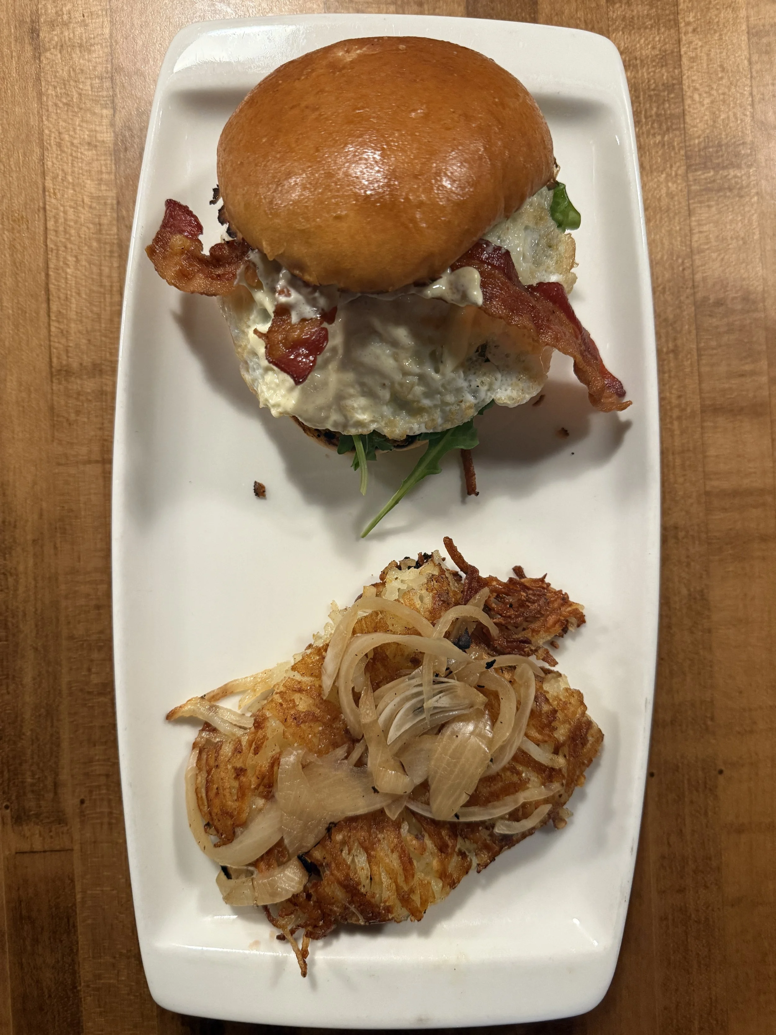 A plate with a bacon, egg, and cheese breakfast sandwich with a brioche bun, fried egg, bacon, and greens, alongside a piece of fish topped with cooked onions.
