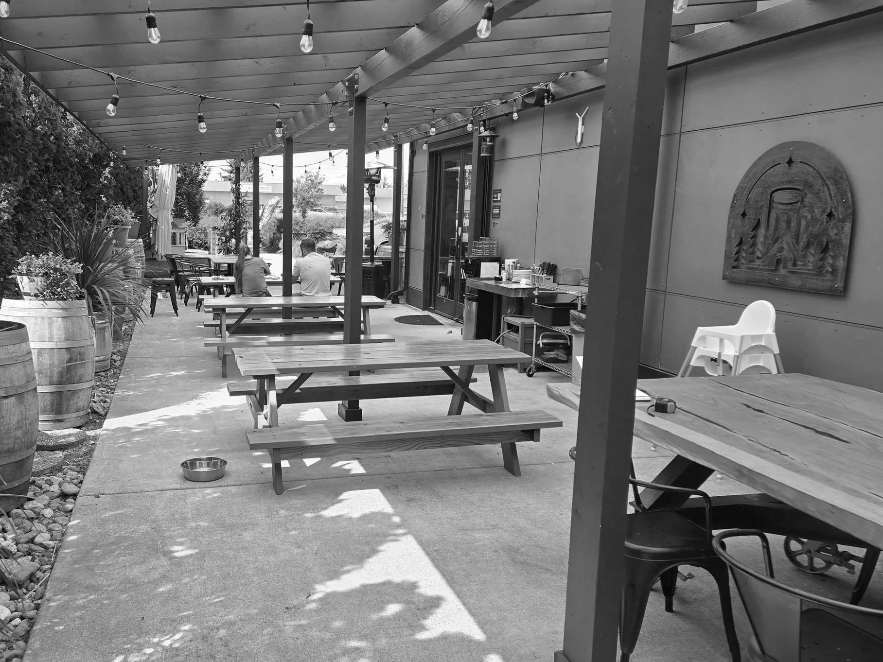 A woman sitting at a wooden table outside a cafe, looking at her phone. She is wearing a hat and glasses, with a large umbrella providing shade. Potted plants and trees surround the area.