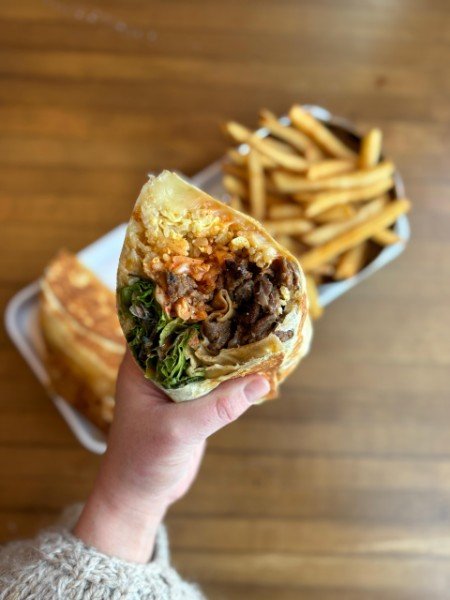 Person holding a half-eaten burger with fries in the background on a wooden table.