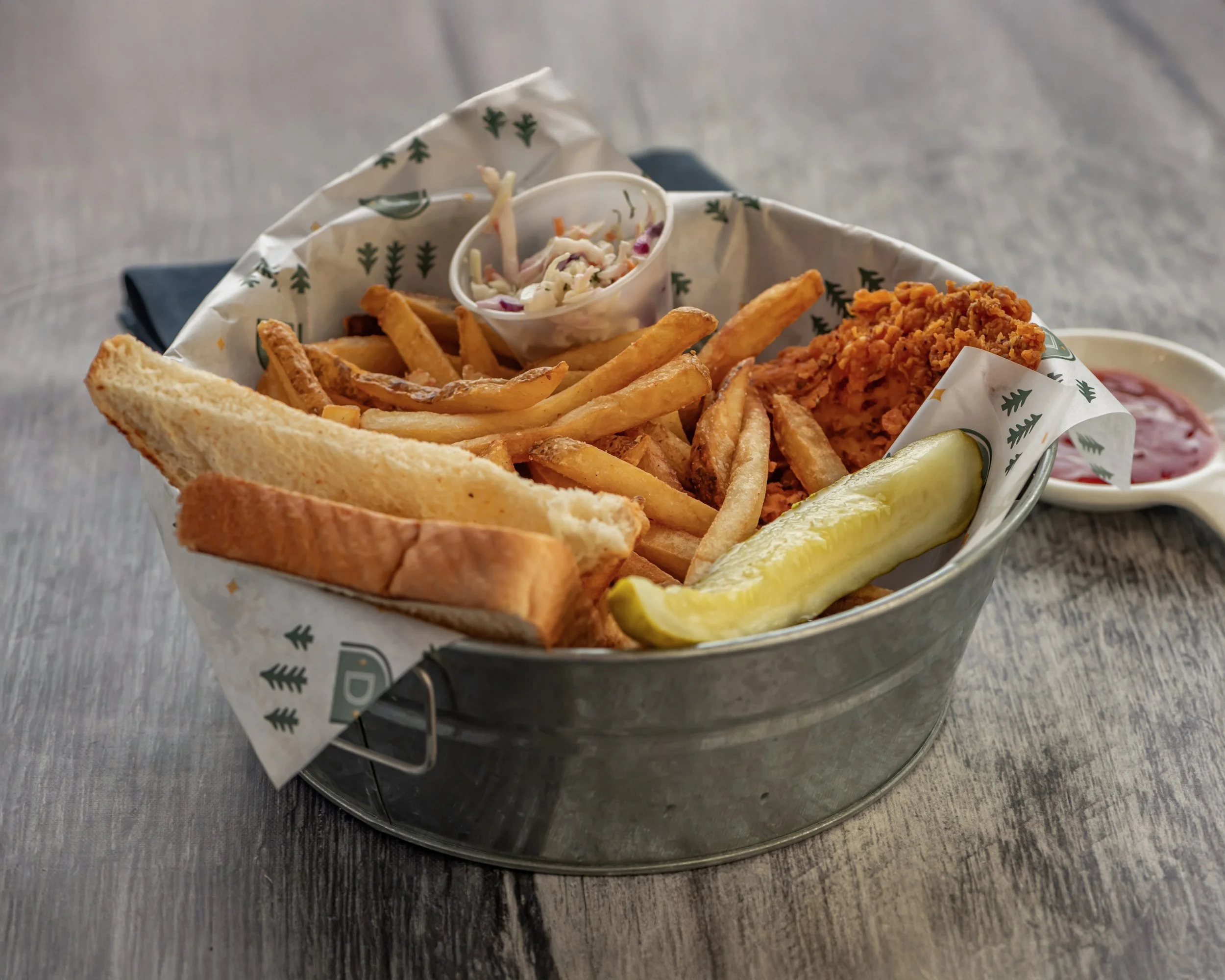 Fried chicken, French fries, coleslaw, toast, pickle, and a side of ketchup in a small white dish, all served in a metal basket lined with paper with a pine tree pattern.