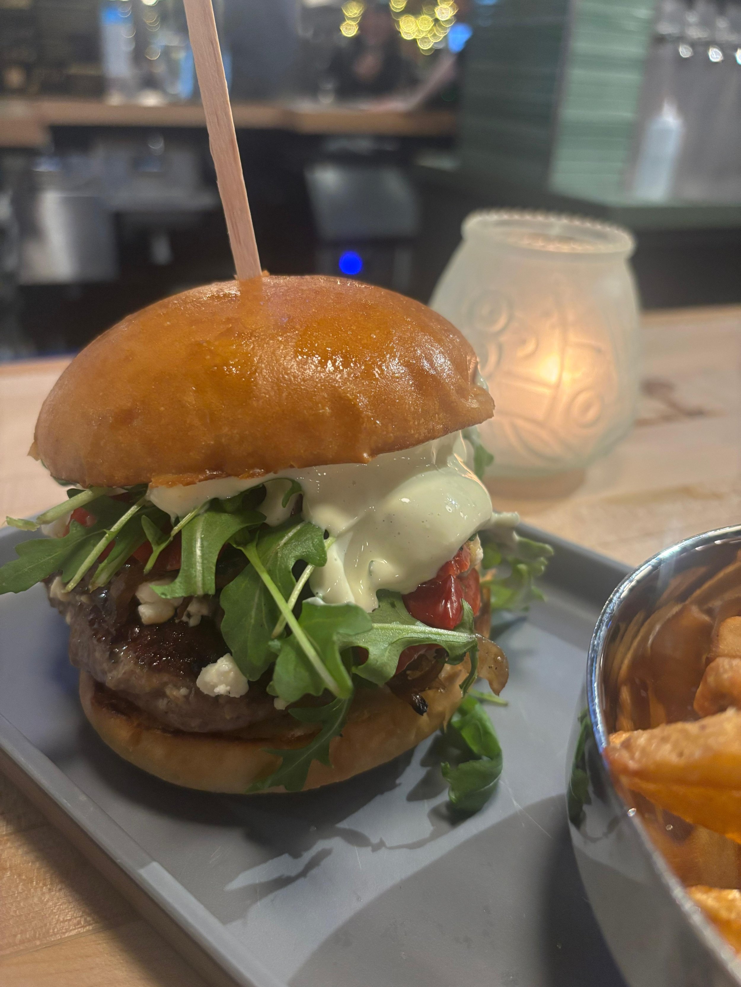 Close-up of a gourmet burger with arugula, cream sauce, tomato, and a beef patty in a brioche bun, served on a white tray with a side of chips, in a cozy restaurant setting.