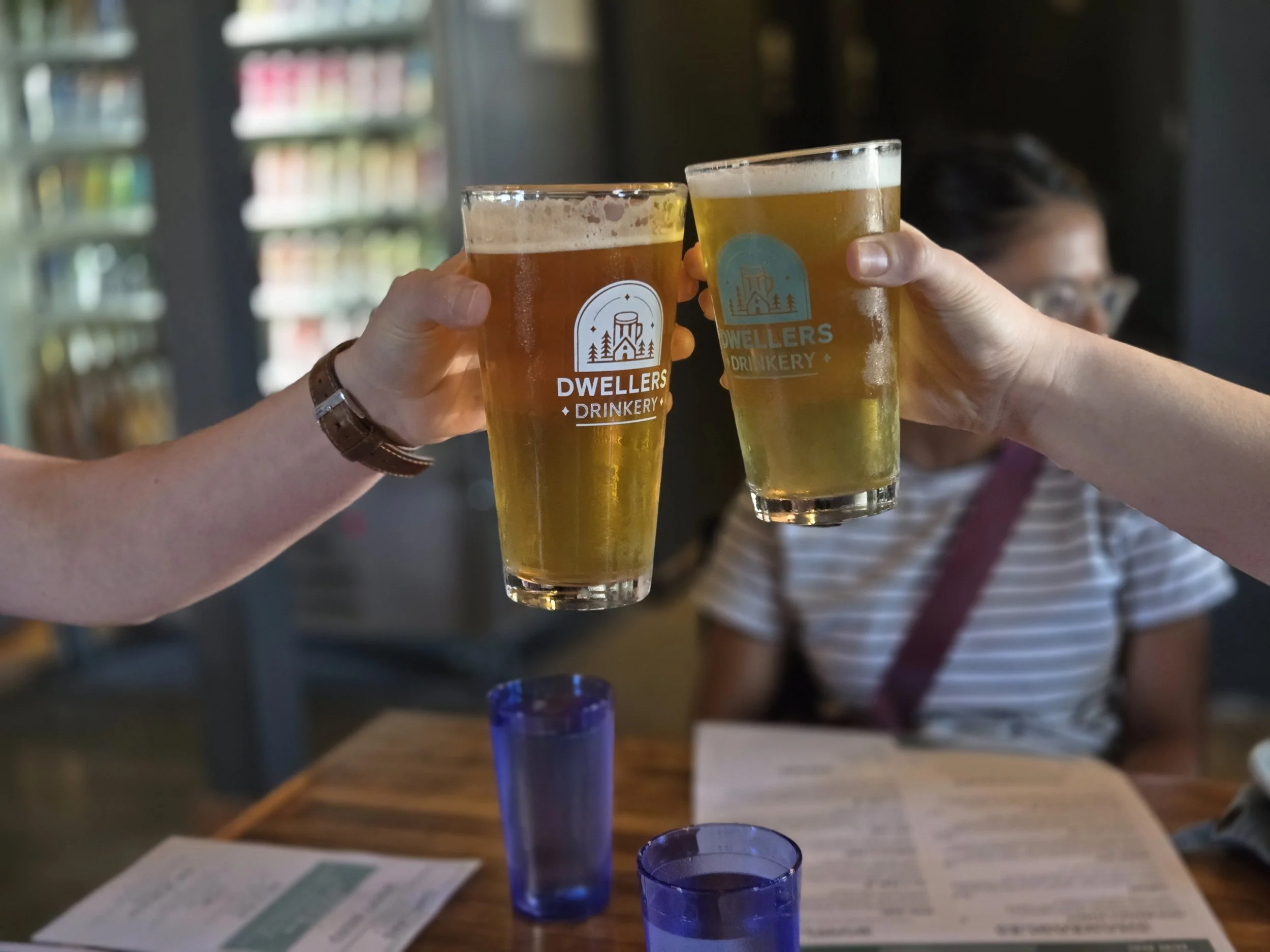 Two people clinking glasses of beer in a bar or restaurant. The glasses have the logo 'Dwellers Drinkery.' In the background, a woman in glasses and a striped shirt is sitting at a wooden table with menus and blue glasses.