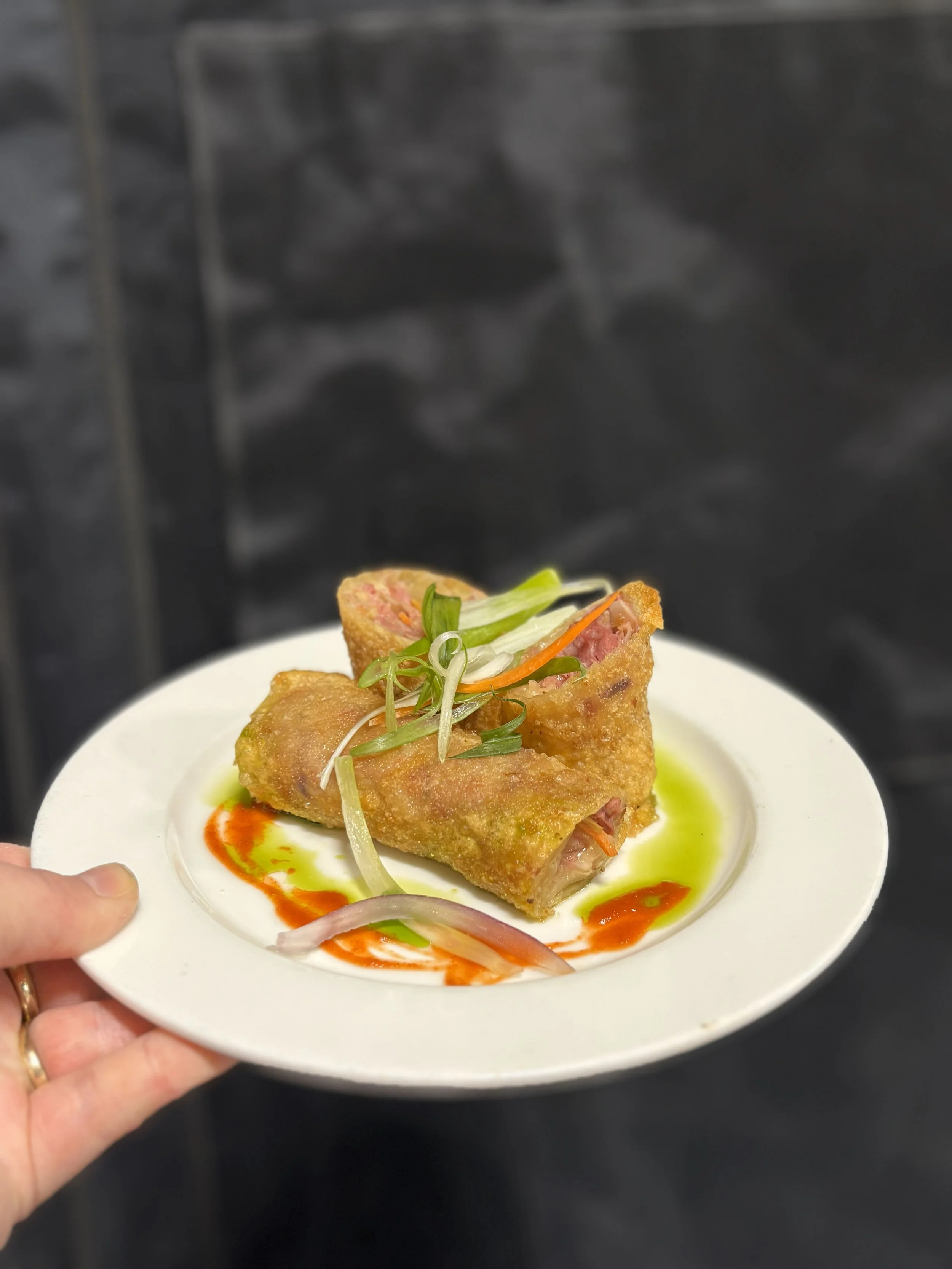 Hand holding a white plate with a dish of fried spring roll garnished with sliced green onions, red and green sauces, and thinly sliced onions.