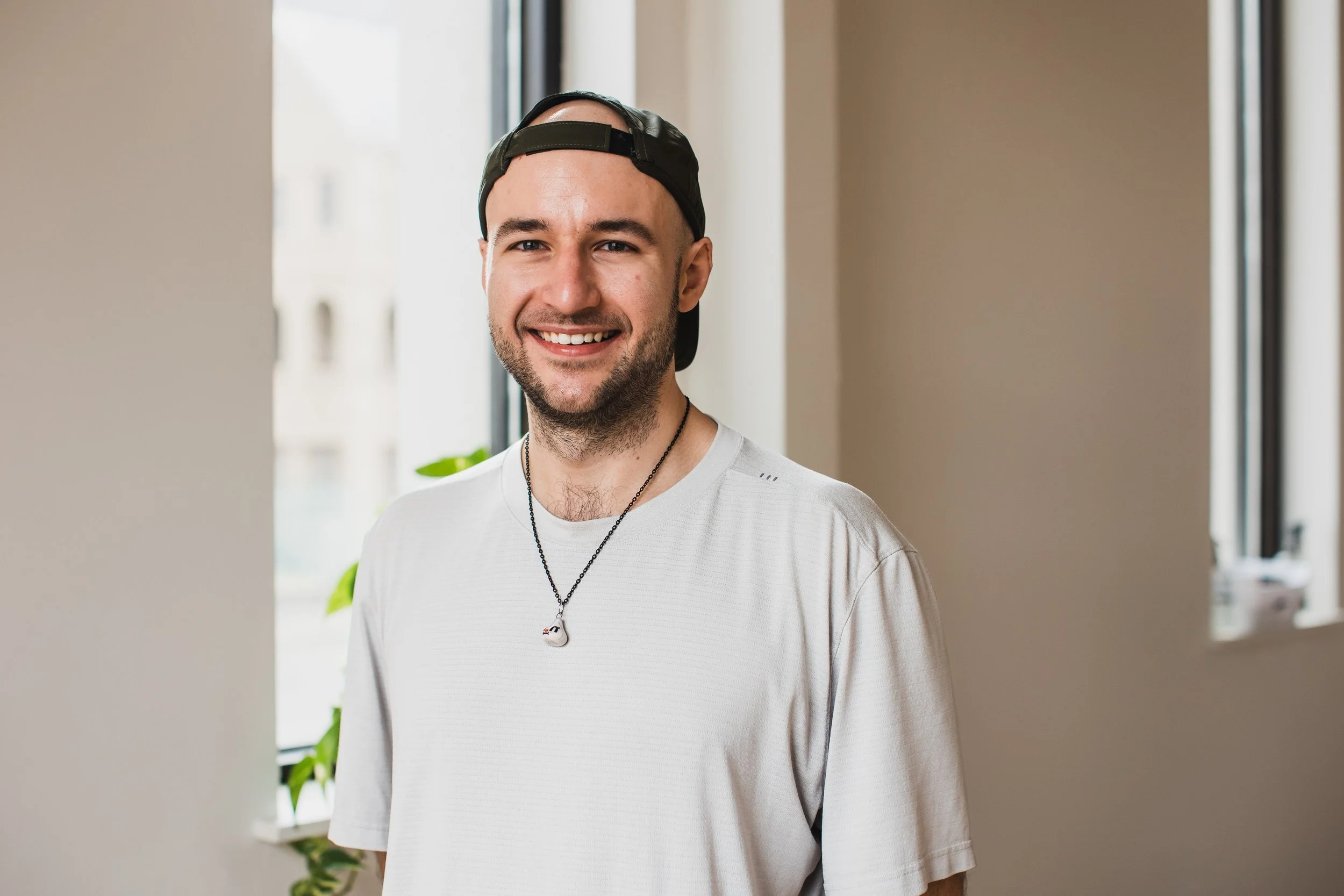 A man with a beard, smiling, sitting cross-legged on a wooden floor in a room with a large leafy plant behind him, a window providing natural light, and a wooden and fabric room divider to the left.