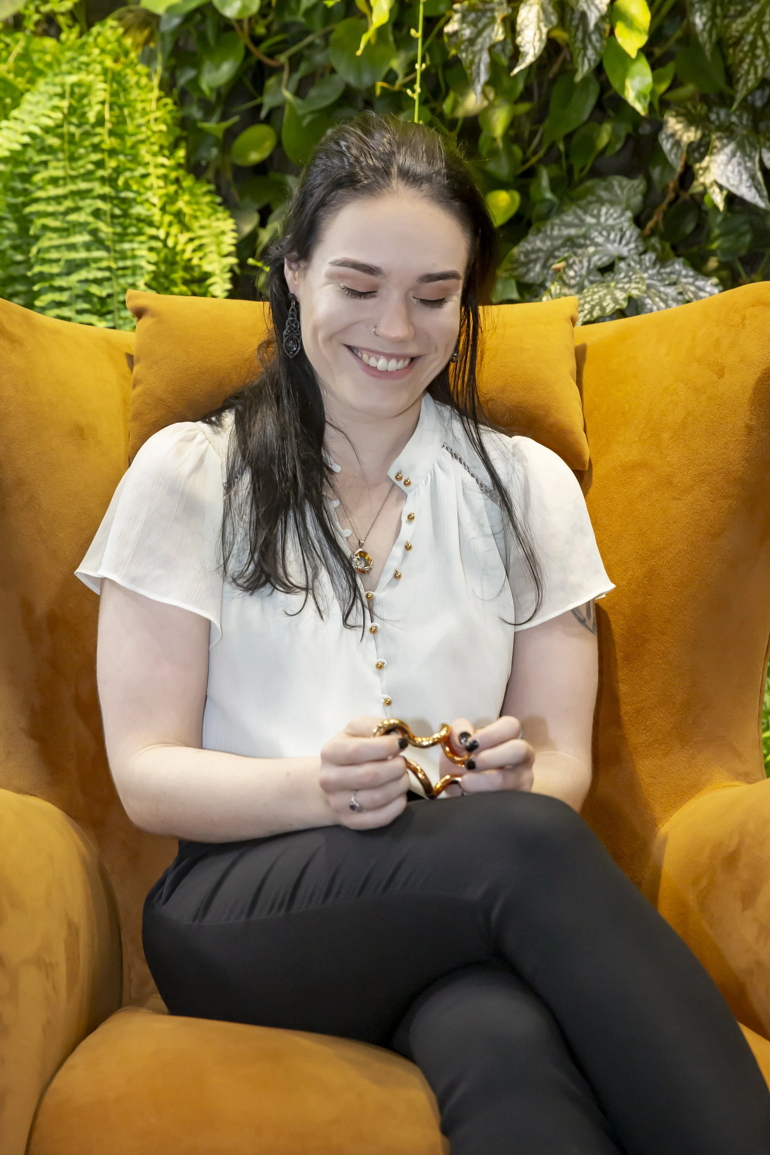 Anna sits smiling on an orange couch. They are holding a gold stimming tool and looking down at it.