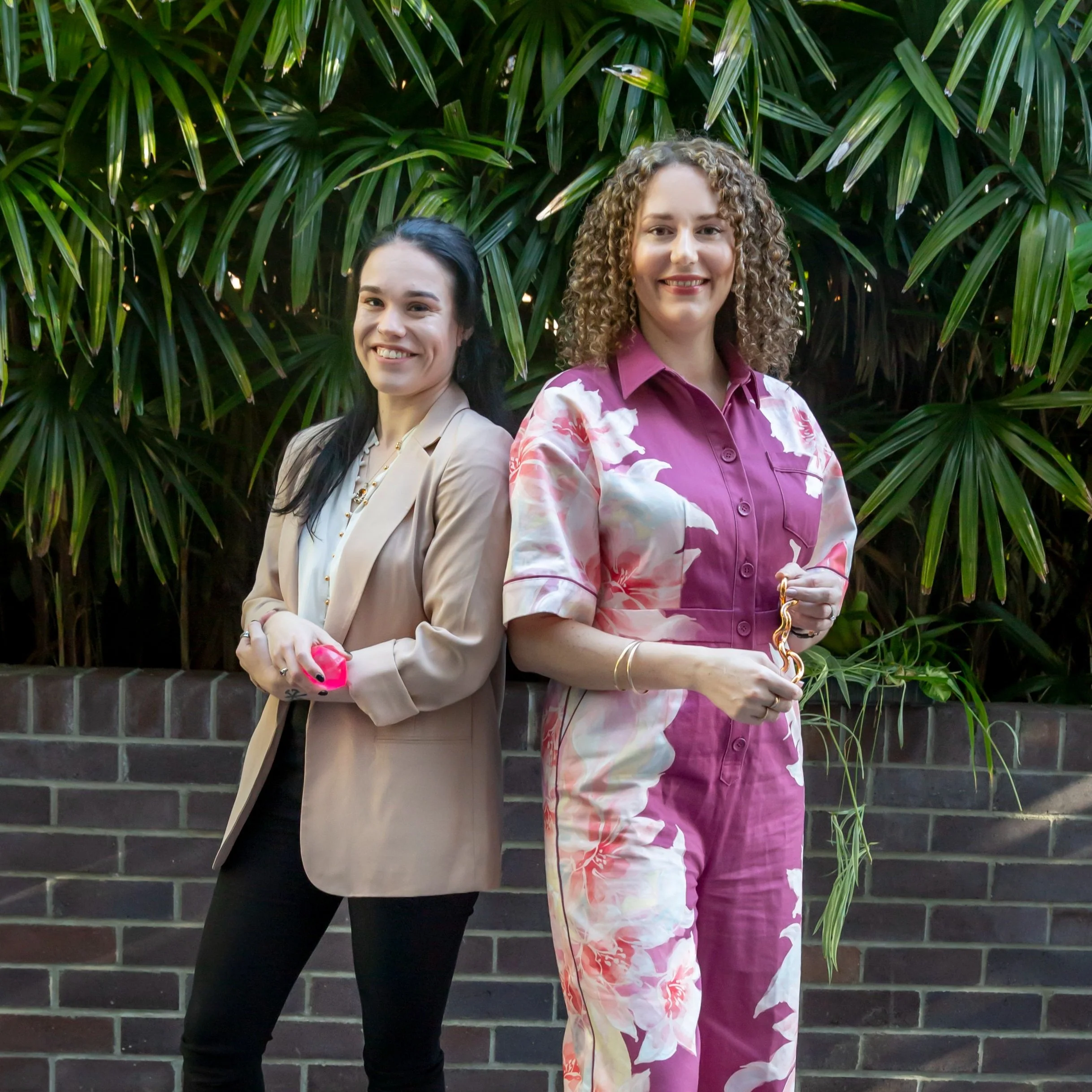 Psychologists Anna and Monique stand back to back in front of a green foliage background. Both are smiling and holding stimming tools.