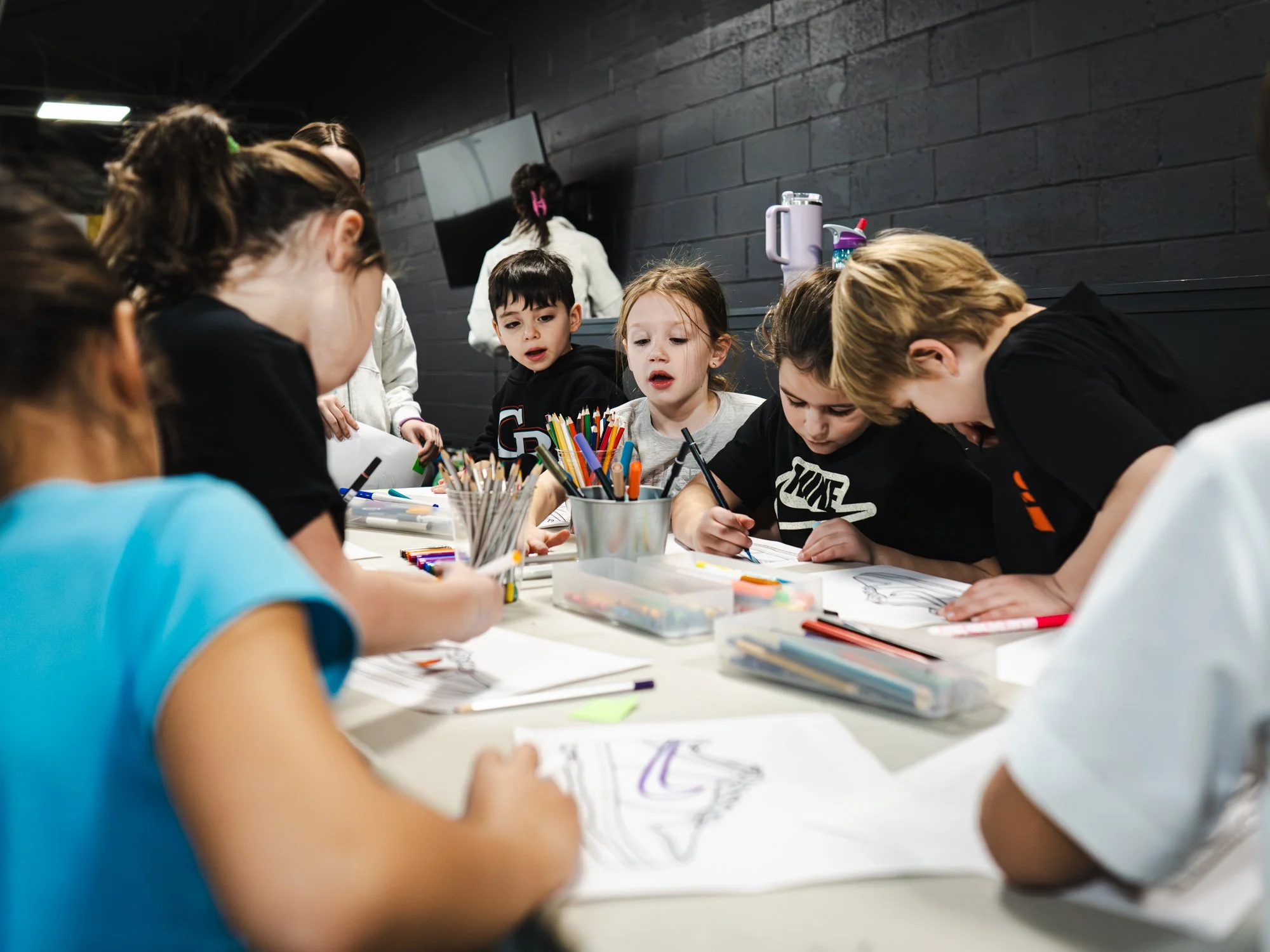 Children gathered around a table engaged in drawing or coloring activities, with various colored markers and supplies on the table.