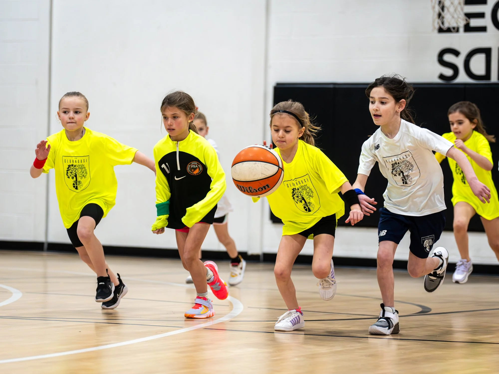 Young children playing basketball in an indoor gym, running together while chasing the ball.