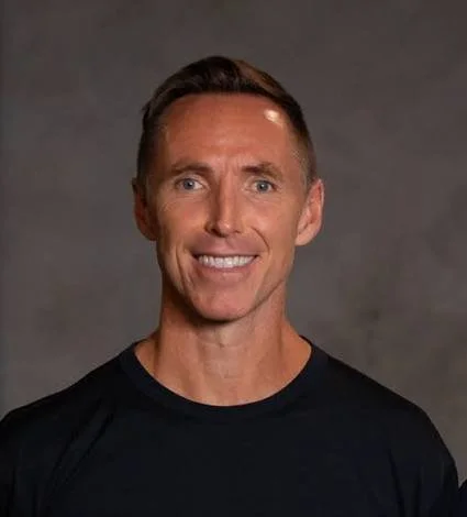 A smiling man with short brown hair wearing a black shirt against a gray background.