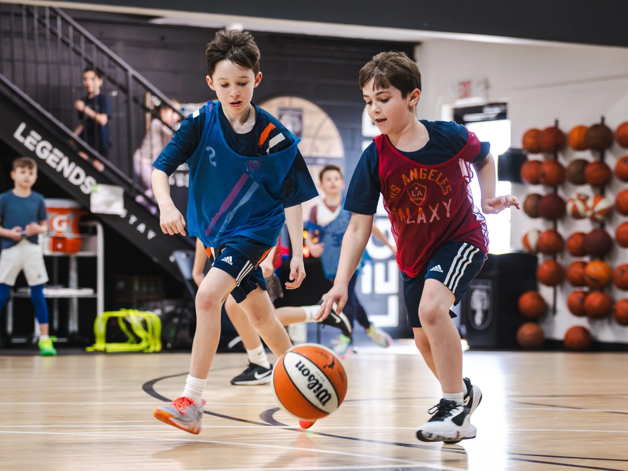 Two young boys playing basketball inside a gym, wearing sports uniforms, with one controlling the basketball and the other attempting to play defense.