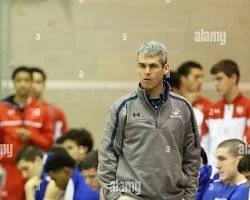 A man with gray hair in a gray jacket standing in front of a group of young athletes. The athletes are wearing sports uniforms, some red and some blue, in an indoor sports facility.