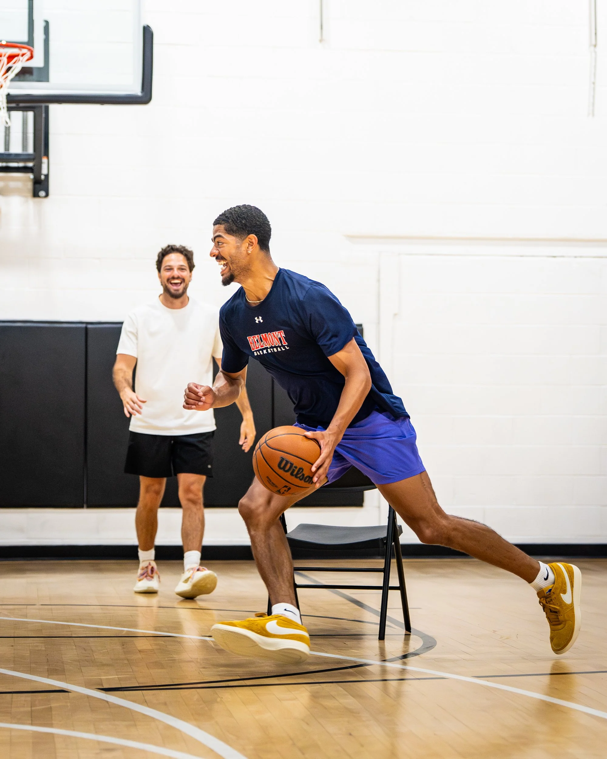 Two men playing basketball in an indoor gym. One man is dribbling the ball and wearing a Belmont basketball shirt, while the other man in a white shirt laughs and stands behind him.