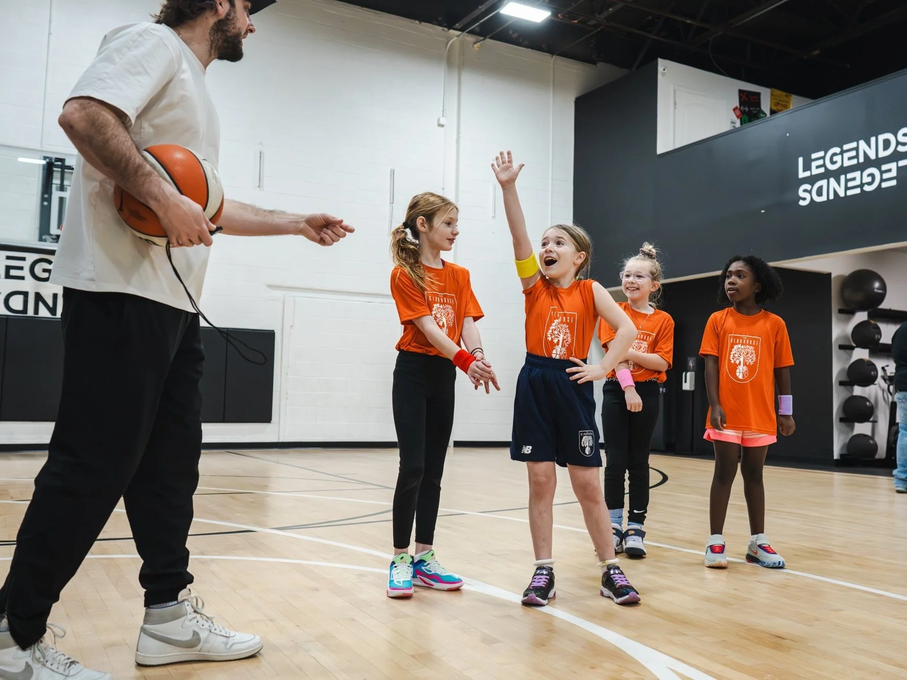A basketball coach teaching young girls in a gym. The coach is holding a basketball, and one girl with a yellow wristband is raising her hand to ask or answer a question. The girls are wearing orange t-shirts and athletic shoes, and they are standing