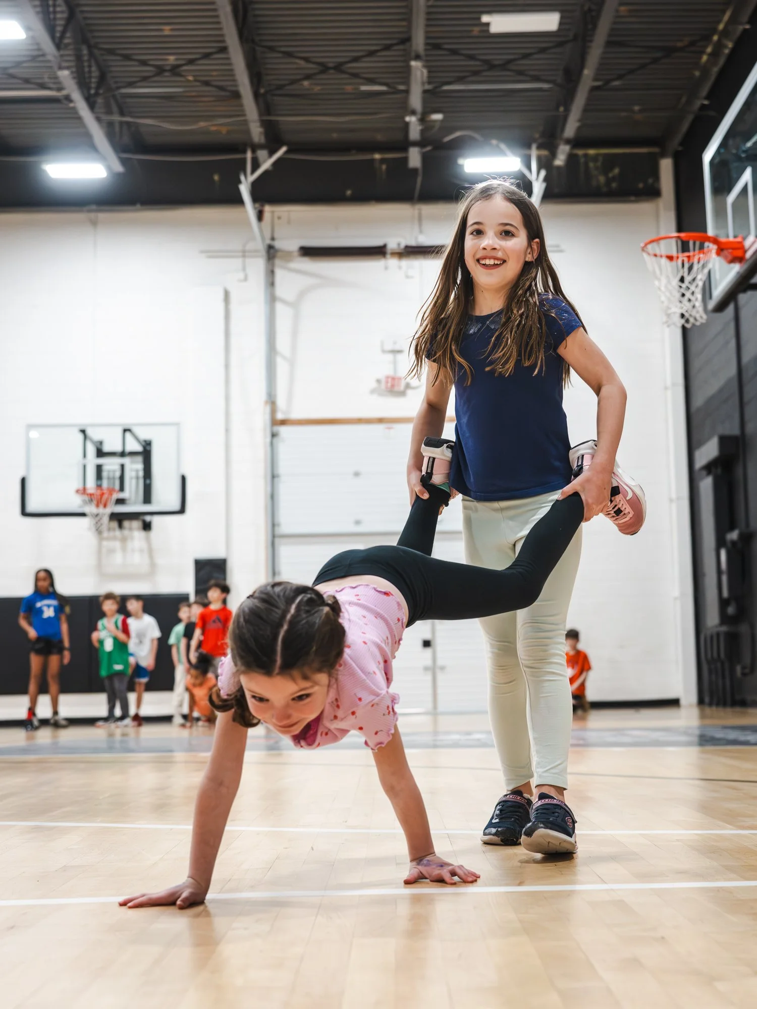 A young girl in a pink shirt is crawling on the gymnasium floor, with a woman holding her legs. The woman has long brown hair and is smiling, wearing a blue shirt and beige pants. In the background, children and adults are standing in a gymnasium wit