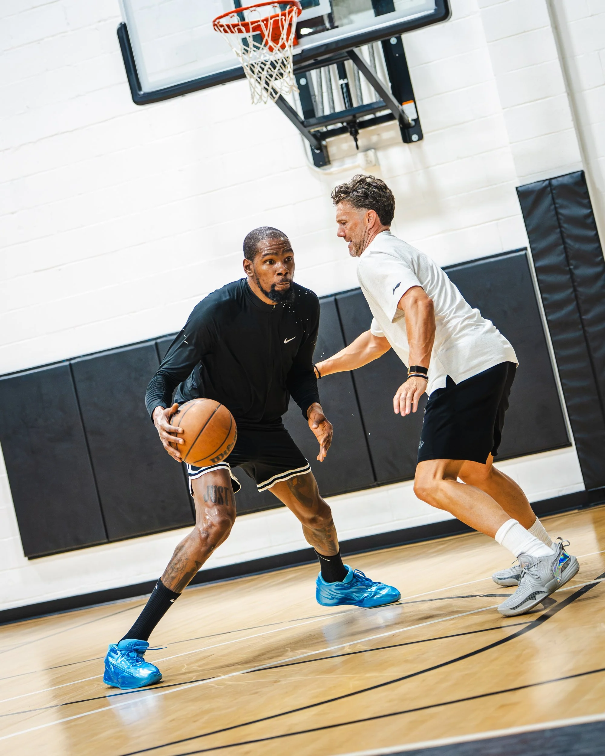 Two men playing basketball on an indoor court with a basketball hoop visible in the background. One man is dribbling while the other is defending him.