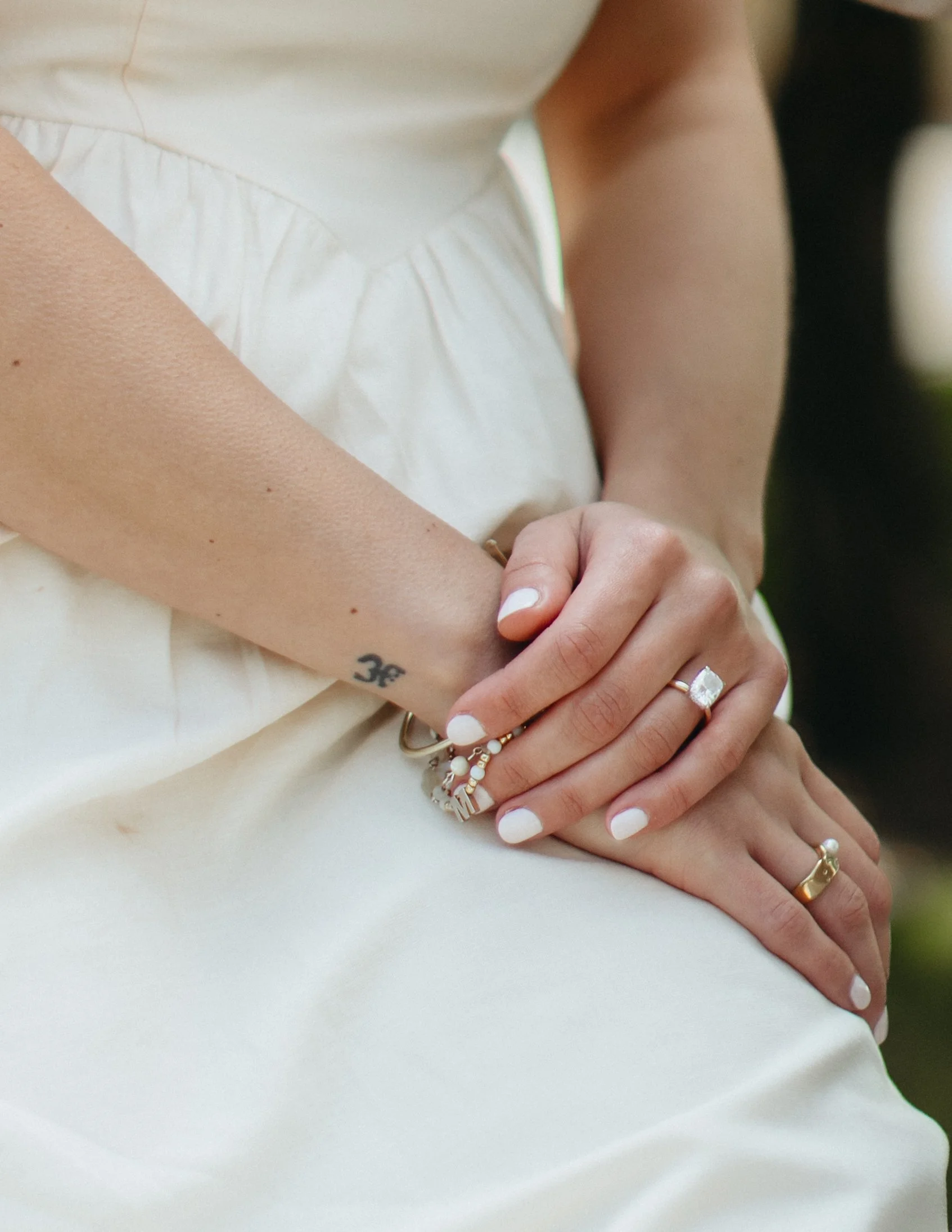 A close-up of a woman's arms crossed in front of her, wearing a white dress, with various rings on her fingers and a tattoo of the Om symbol on her wrist, with a blurred outdoor background.