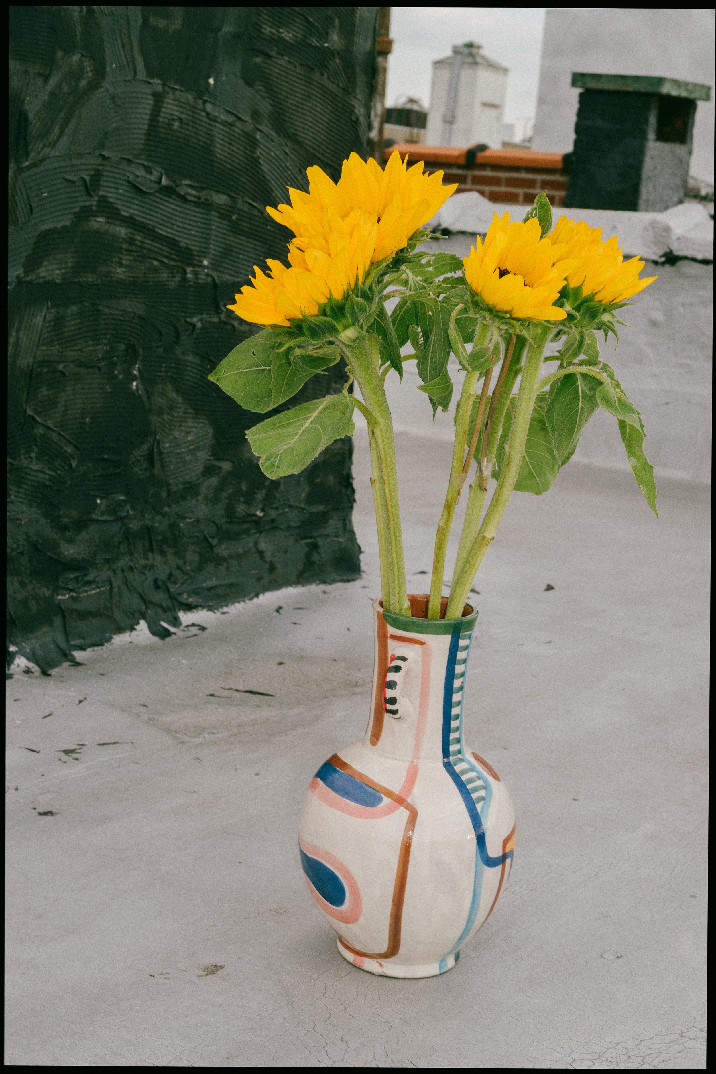 Yellow sunflowers in a colorful, abstract-patterned ceramic vase on a concrete surface with rooftop buildings in the background.