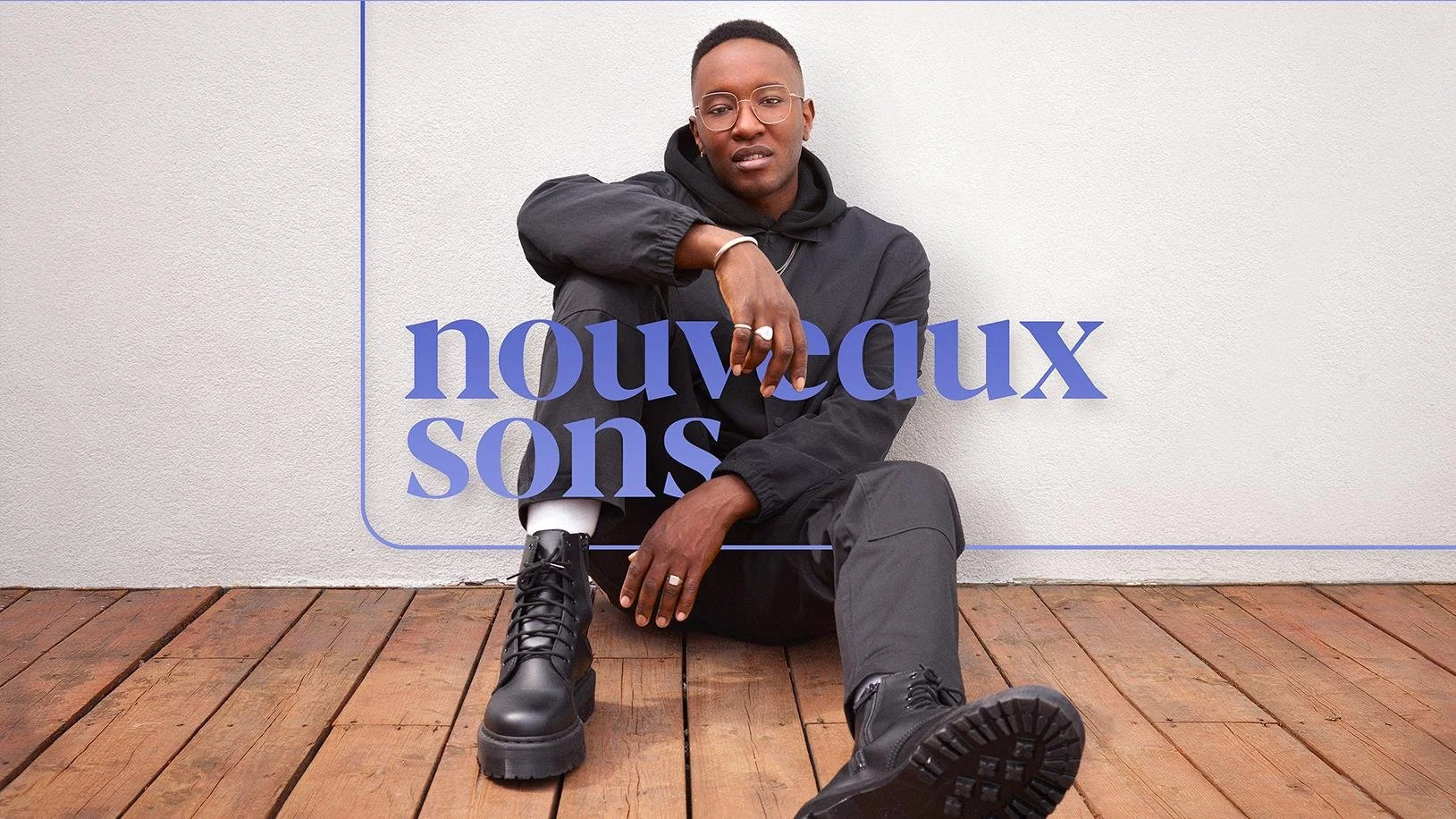A person wearing black clothing, glasses, and black boots sitting on wooden flooring in front of a plain white wall with translucent text overlaid that reads "nouvaux sons."
