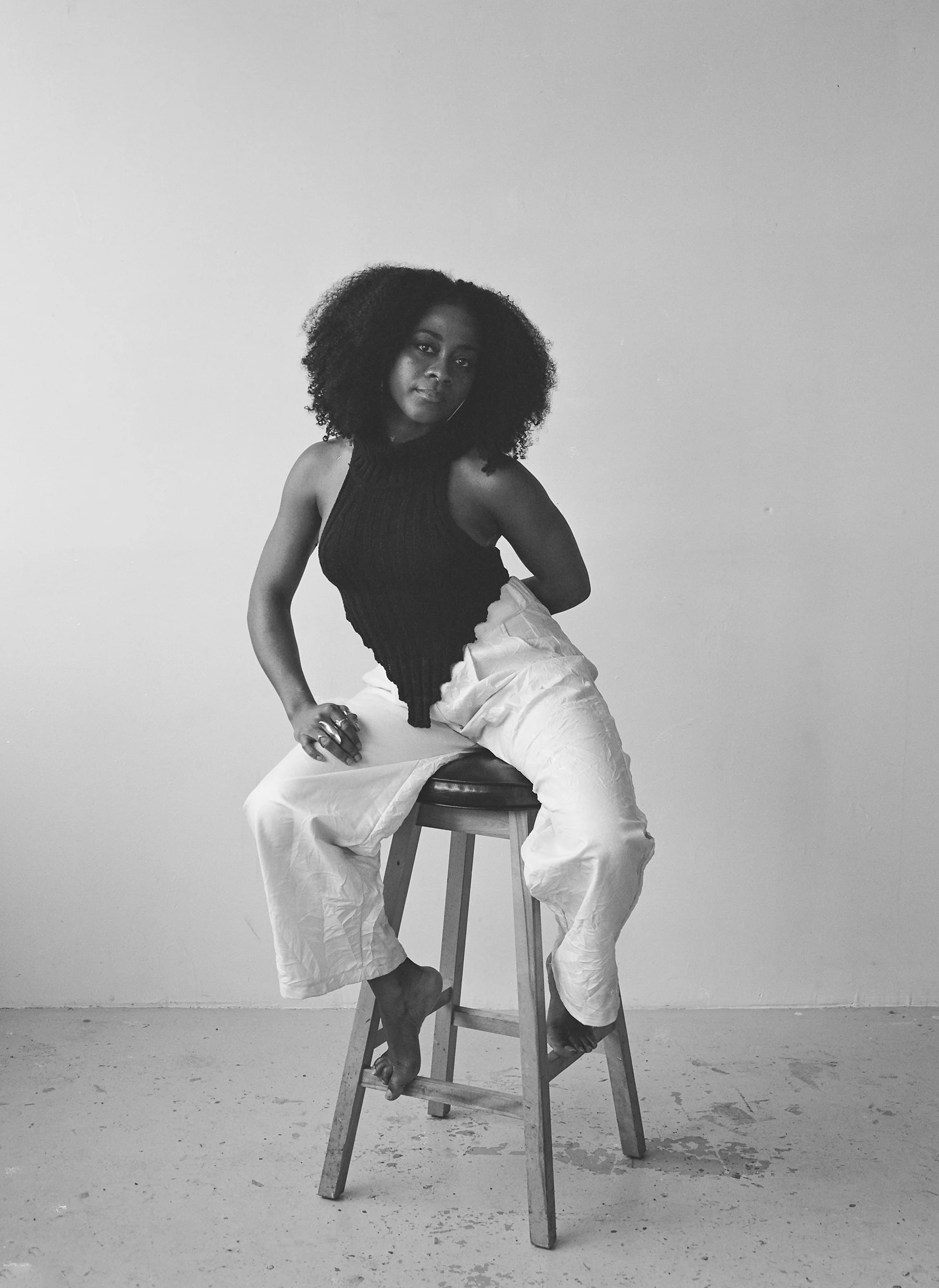 A woman with curly hair sitting on a stool against a plain wall, wearing a sleeveless dark top and light-colored loose pants.