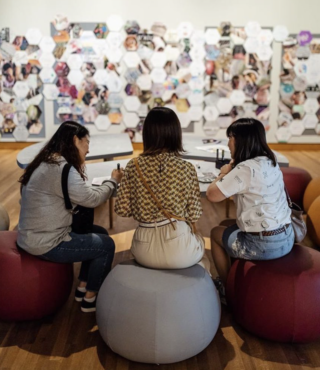 Three women are sitting on chairs and a round ottoman in a room with a wall covered in photos. They are engaged in conversation and one woman is writing in a notebook.