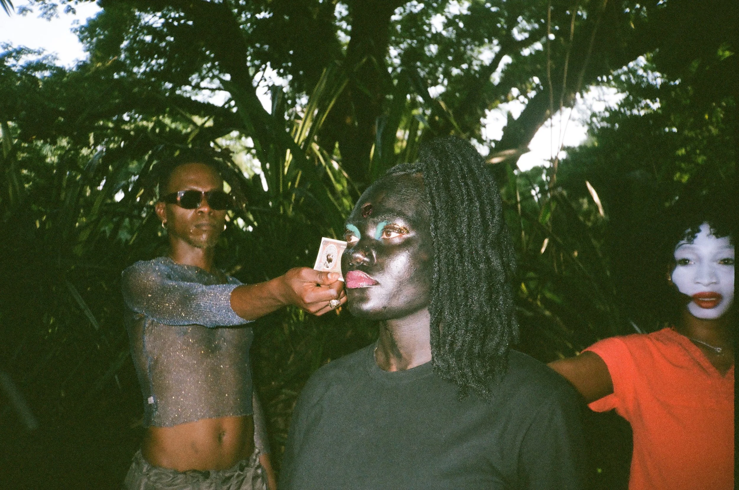 Three people in an outdoor setting with lush green foliage. One person with dark makeup and dreadlocks is receiving a photograph from another individual wearing sunglasses and a glittery top. A third person with painted face in white makeup and red lipstick is smiling on the right.
