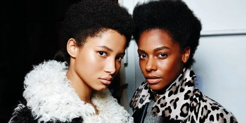 Two women with natural hair posing indoors, one wearing a white fur-lined coat and the other in a leopard-print coat.