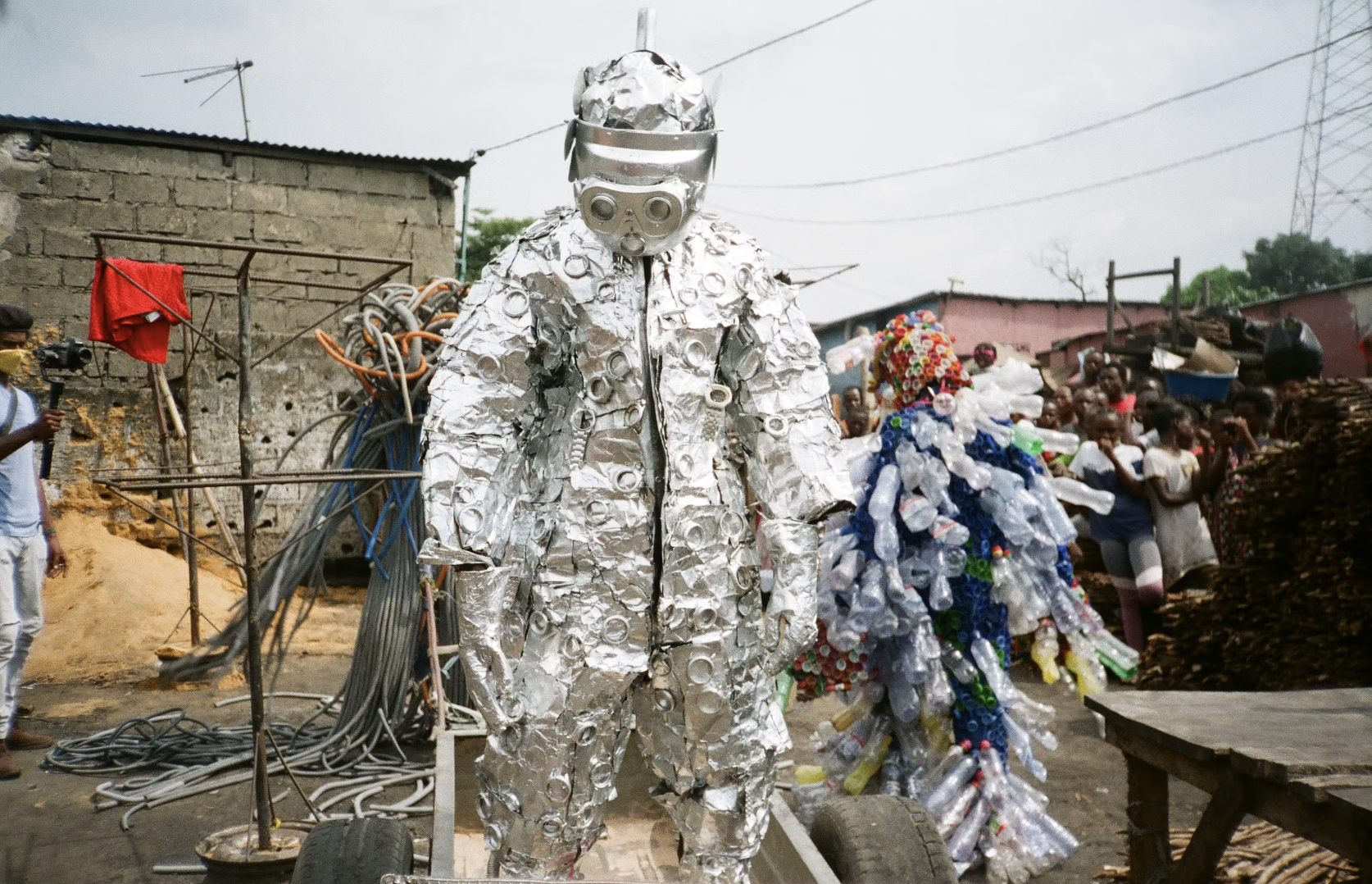 A person dressed in a silver suit made of recycled aluminum cans and a helmet with goggles, standing on a small vehicle in a busy outdoor setting with onlookers and structures in the background.
