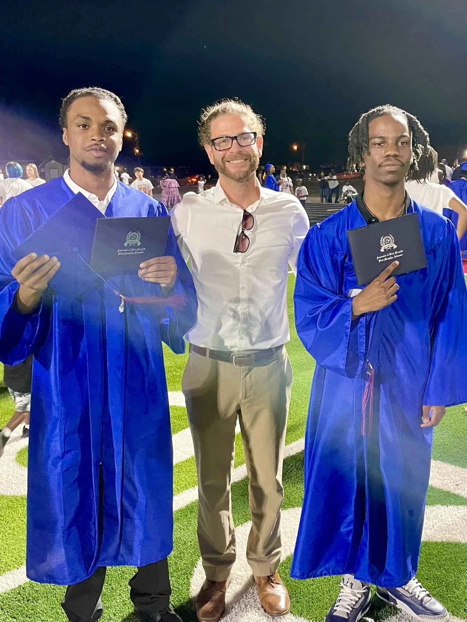 Three men stand together at night on a football field, two wearing blue graduation gowns, one in the middle in casual white shirt and khakis. The two graduates hold diplomas, and the scene is illuminated with bright lights.