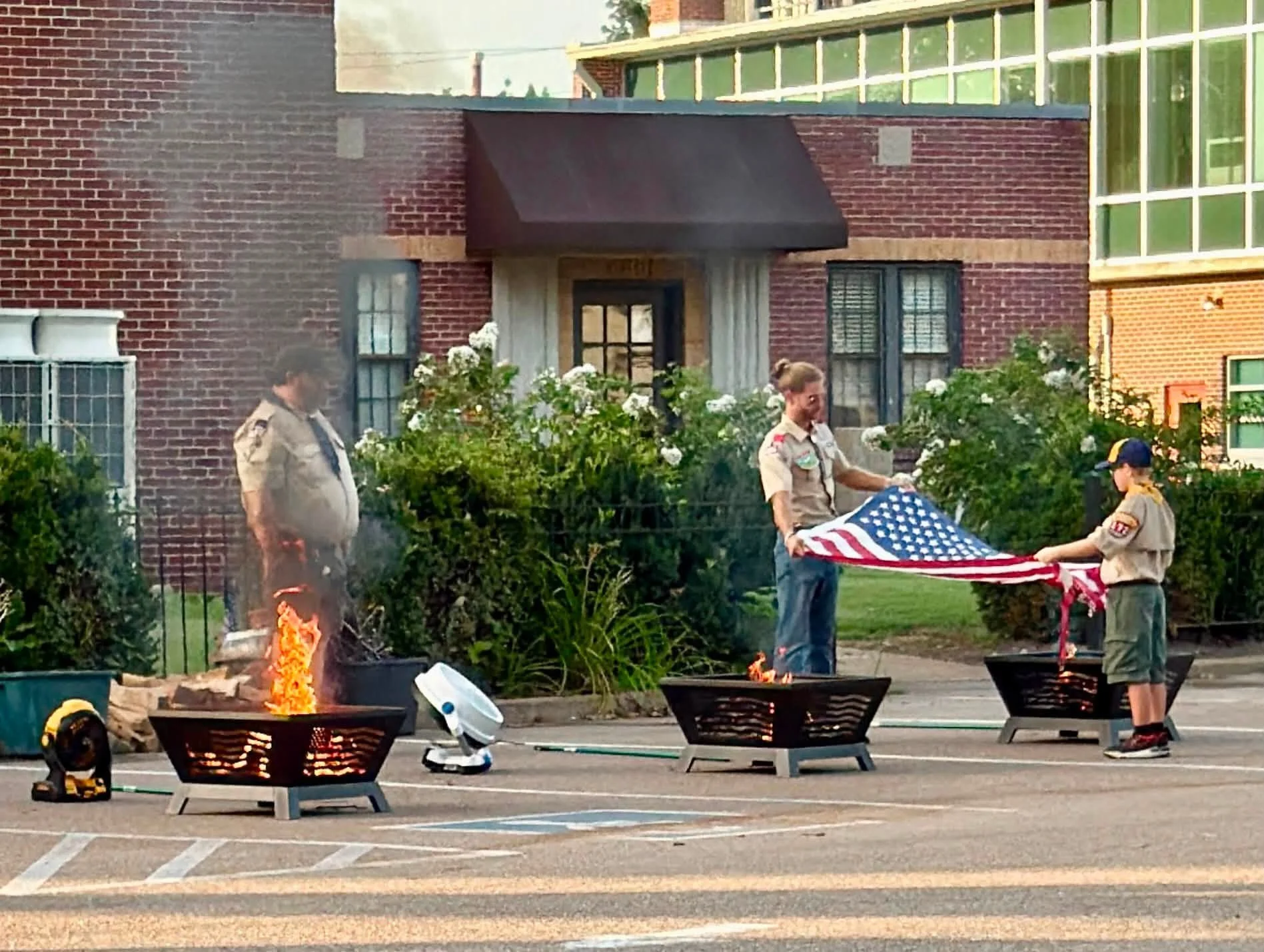 Boy Scouts raising an American flag during a patriotic ceremony, with small fires burning in metal fire pits in front, on a street with brick buildings in the background.