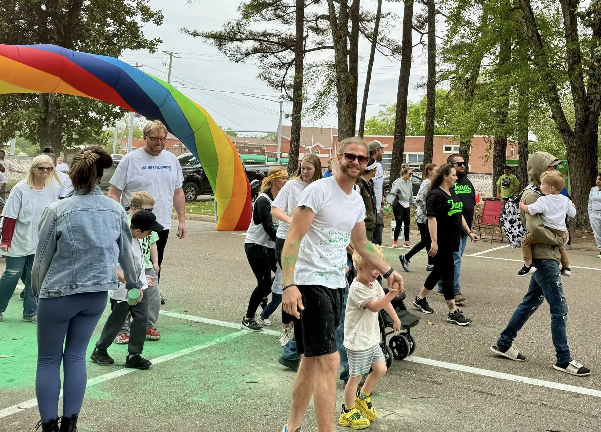 People gathered outdoors under trees, some wearing white T-shirts, participating in a group activity or celebration, with a large rainbow-colored arch in the background.
