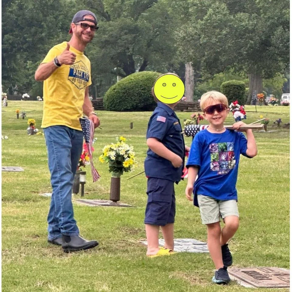 A man giving a thumbs-up next to two children standing on grave markers in a cemetery, with flowers on the graves and trees in the background.