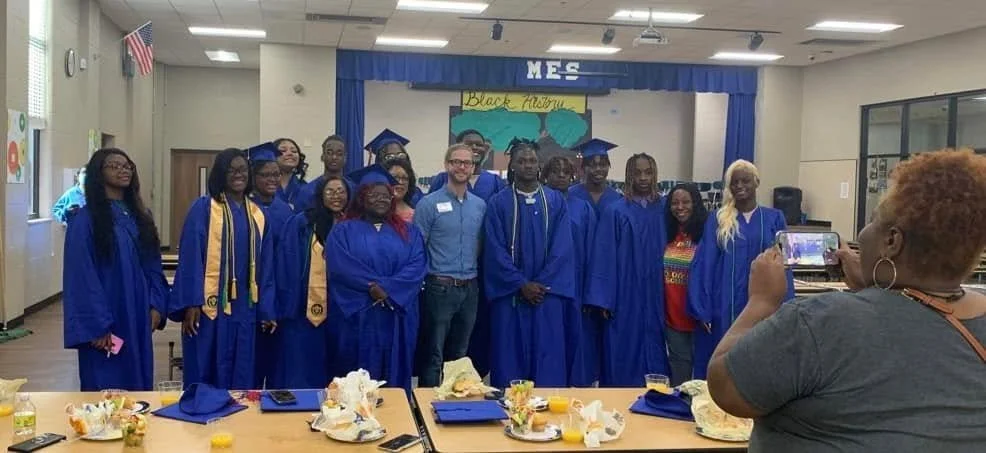 Graduation ceremony with students in blue caps and gowns, standing on stage, celebrating at MES school. A woman is taking a photo of the group.