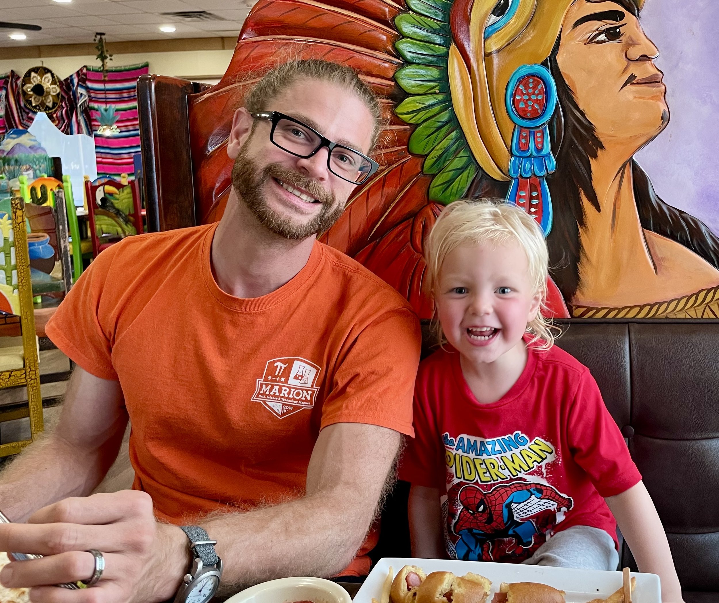 A man with glasses and a beard smiling at the camera next to a young girl with blonde hair and a wide smile, both seated at a table with food. Behind them, there is a colorful carved wooden Native American chief sculpture. The scene appears to be in a restaurant or arcade with bright and festive decor.