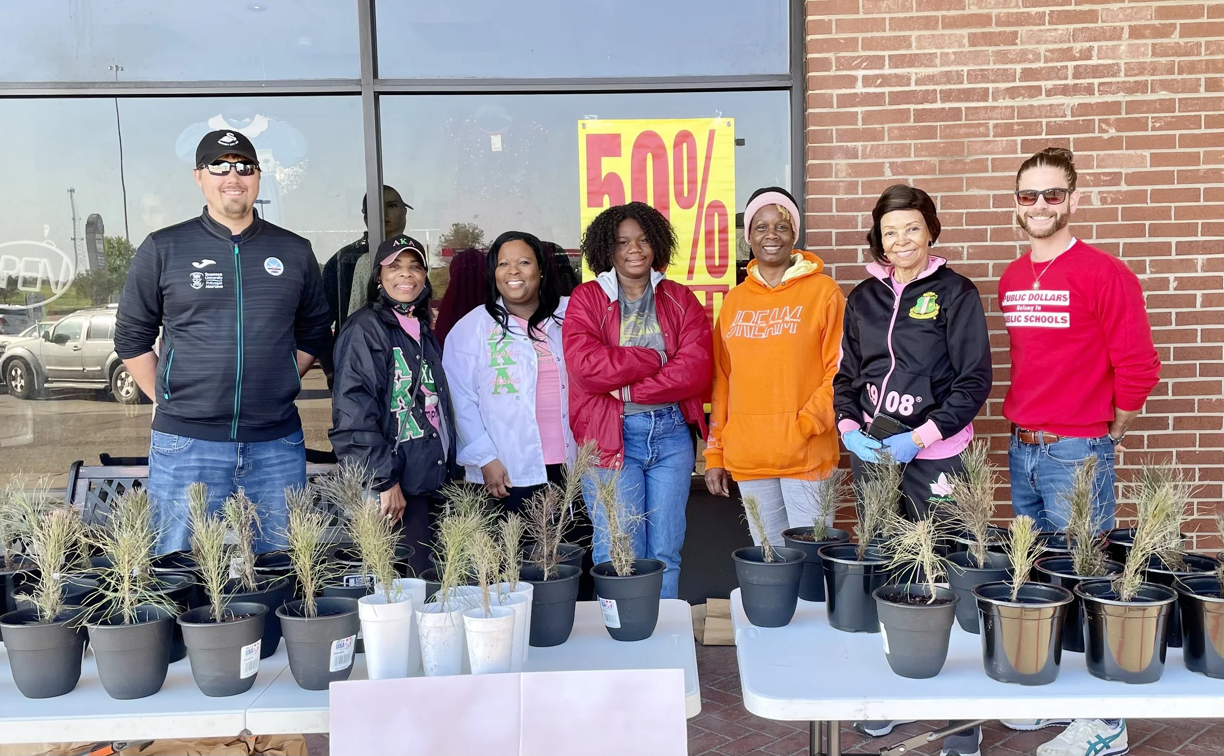 Group of eight people standing behind a table with small potted plants for sale or display outside a building with a large yellow sign showing '50%'. They are smiling and dressed casually, some wearing jackets or hoodies, with a brick wall and a wind