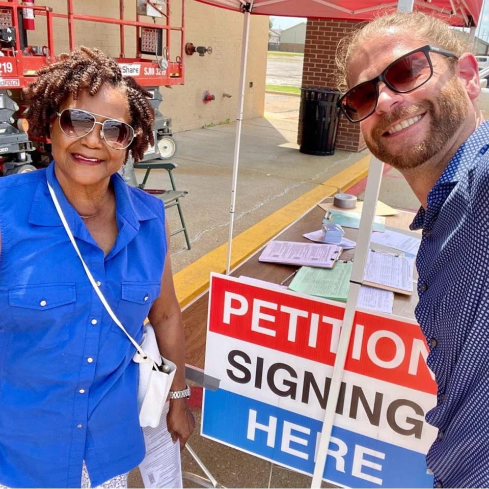 A woman in a blue sleeveless shirt and sunglasses and a man in a blue patterned shirt and sunglasses smiling at a petition signing table under a red canopy outside a building.