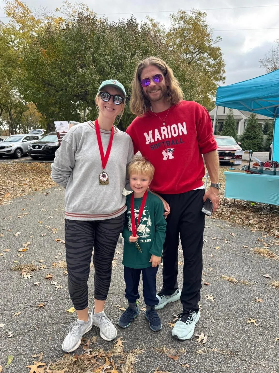 A woman, man, and young boy standing outside during fall with trees and parked cars in the background. The woman and boy are wearing medals on red ribbons, and everyone is dressed casually with the woman and man wearing sunglasses.