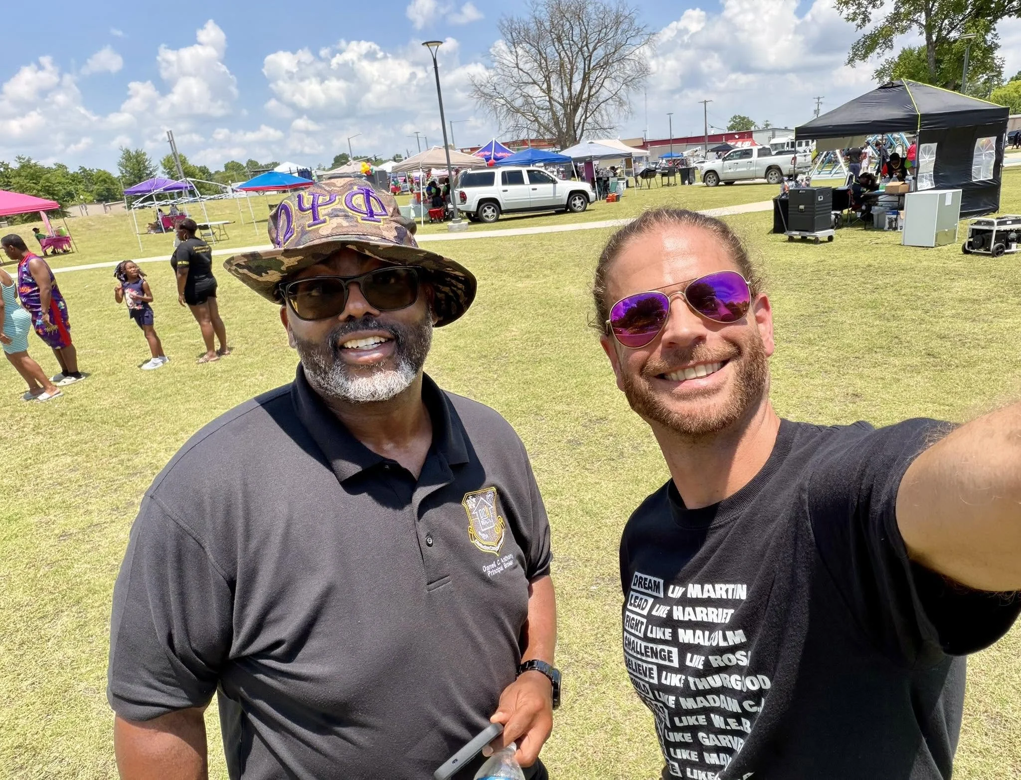 Two men smiling in the foreground at an outdoor event with tents, vehicles, and people in the background under a partly cloudy sky.