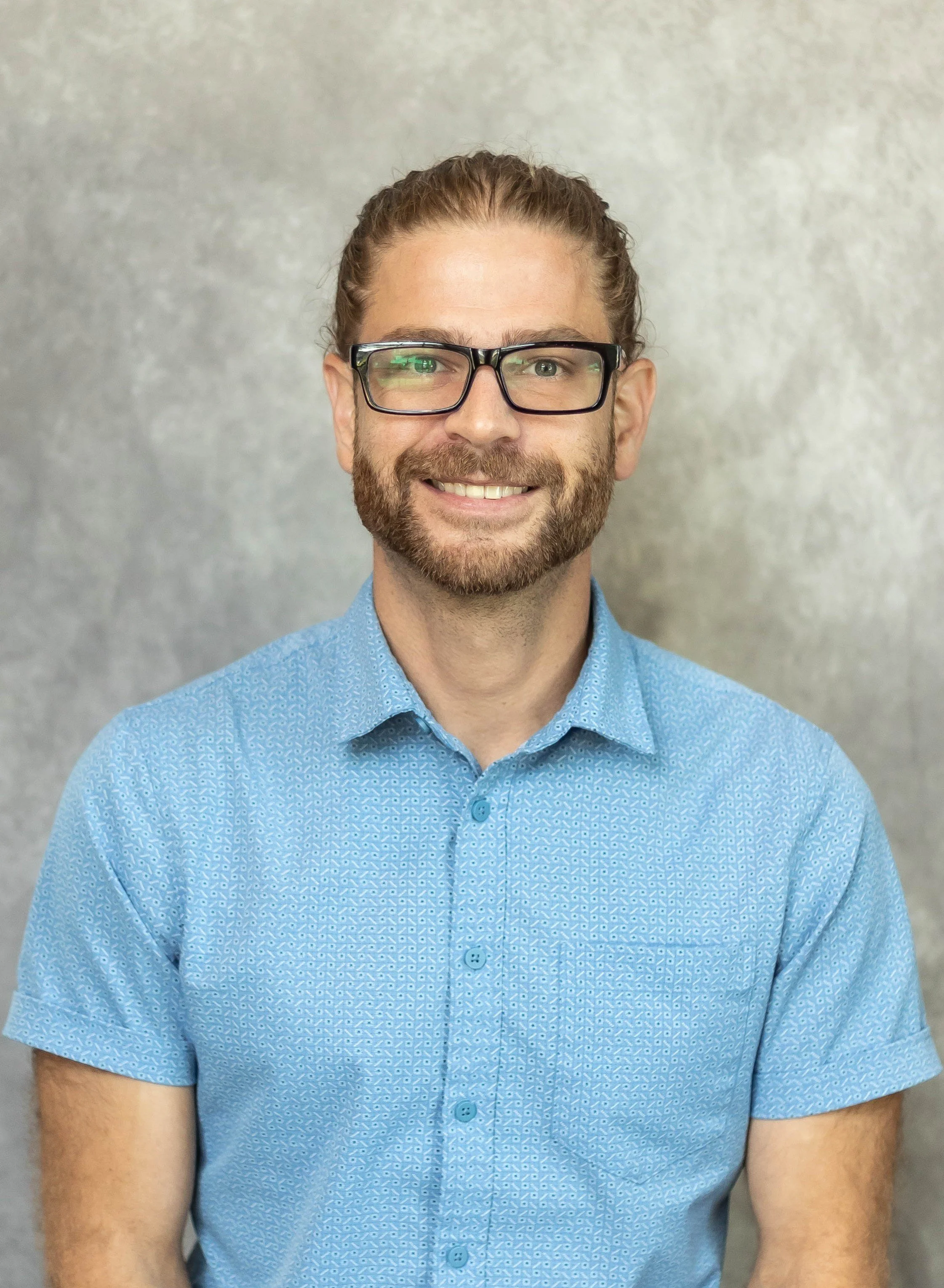 A smiling man with glasses, a beard, and long hair tied back, wearing a light blue patterned shirt, standing against a plain gray background.