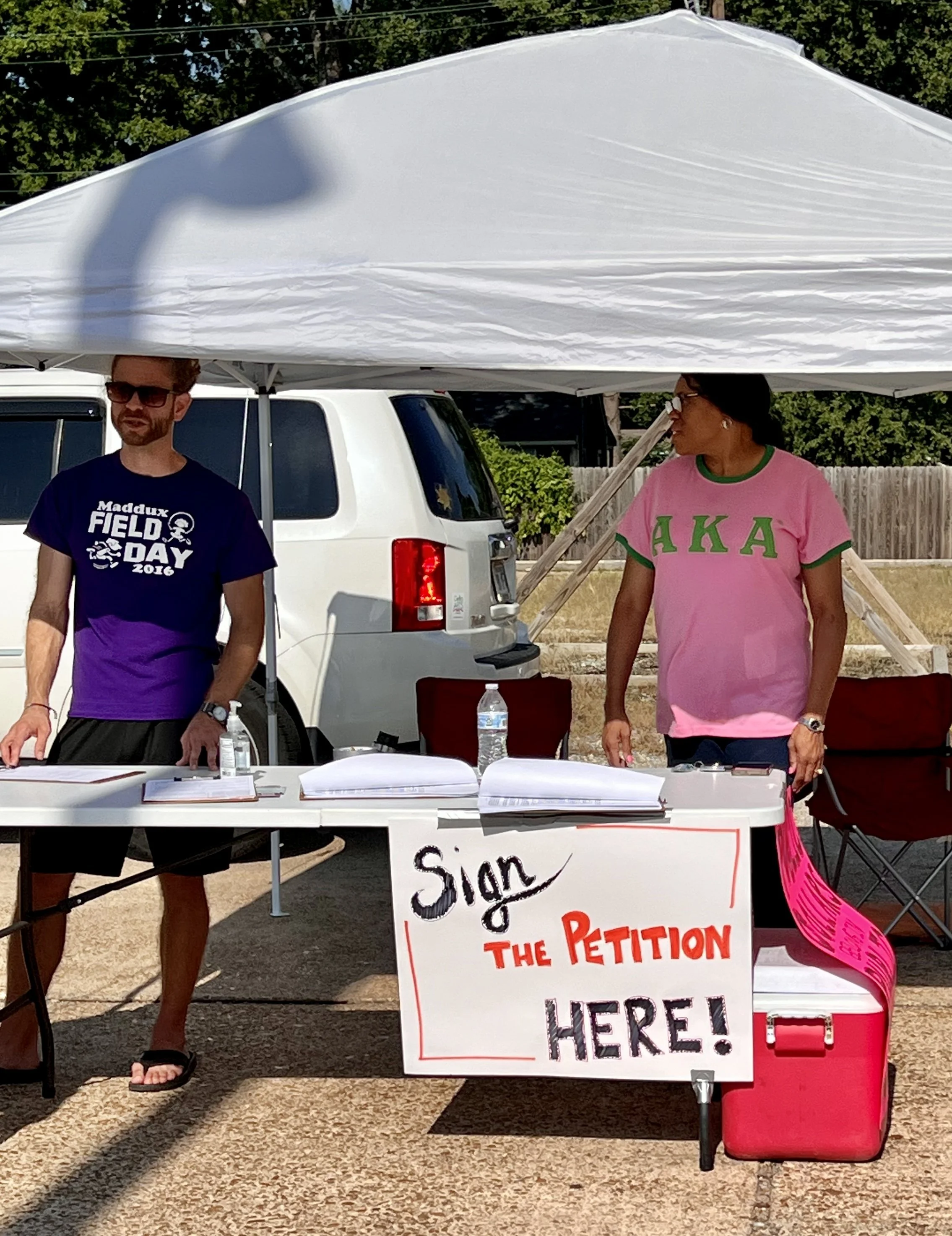 A woman and a man standing behind a table at a petition signing event outdoors, under a white canopy tent. The table has sign-up papers, hand sanitizer, and water bottles. The woman is wearing a pink shirt with green Greek letters and the man is wear