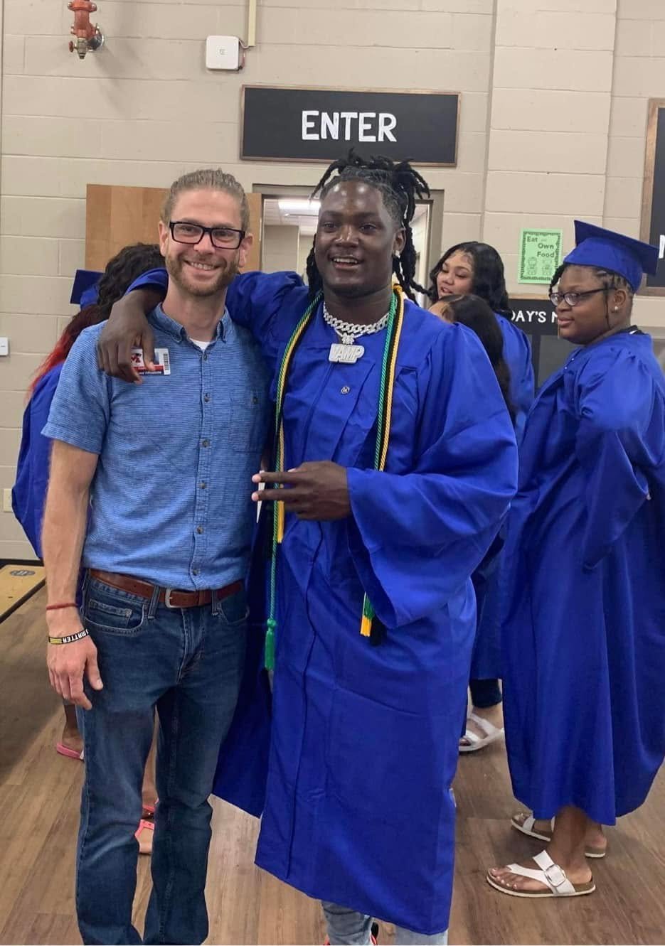 Two men, one in a blue shirt and jeans and the other in a blue graduation gown, stand together smiling at a graduation ceremony, with others in blue graduation gowns in the background.