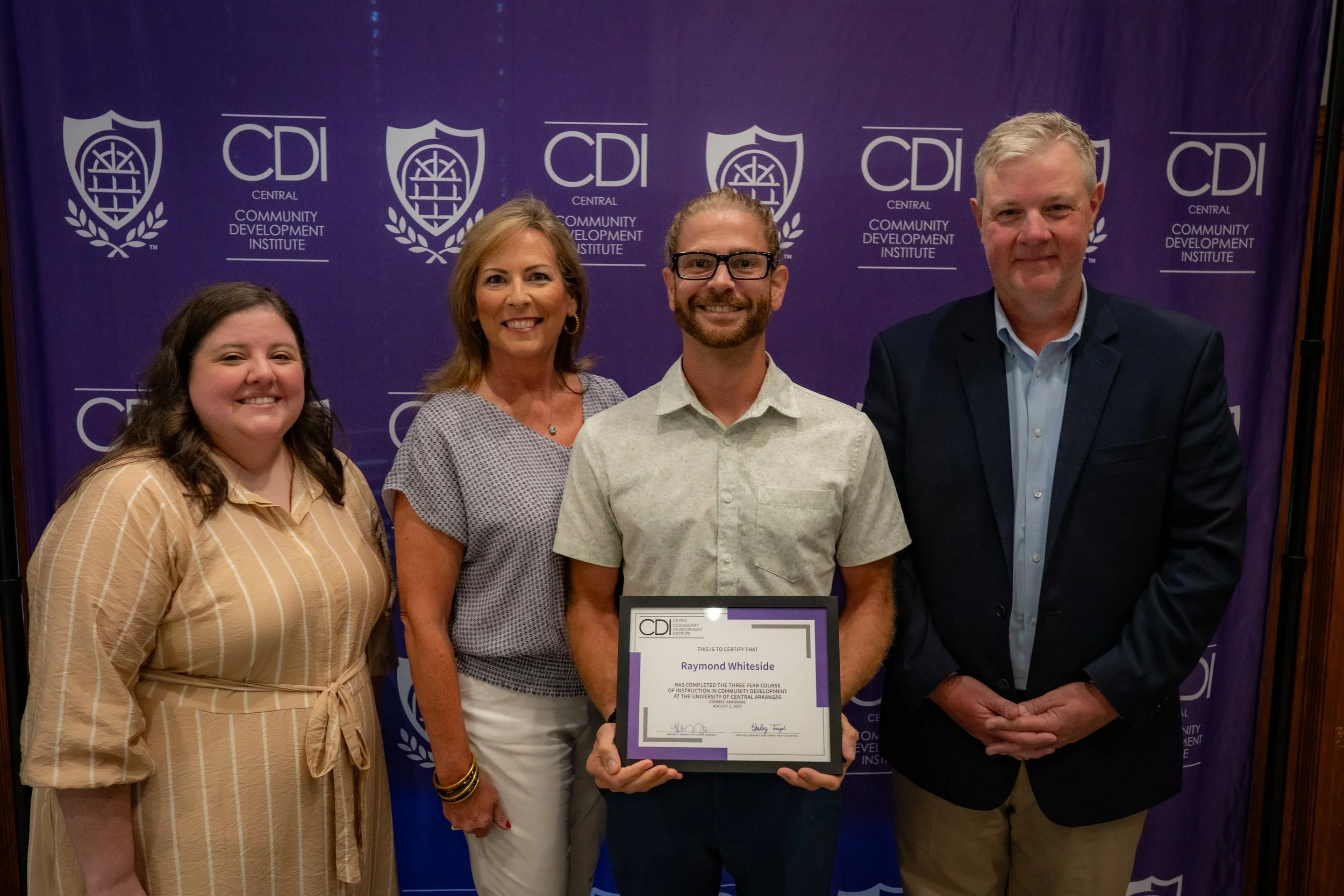 Group of five people posing in front of a purple backdrop with the logo and name of the Central Community Development Institute. One woman in the center holds a certificate for Raymon Whiteside.