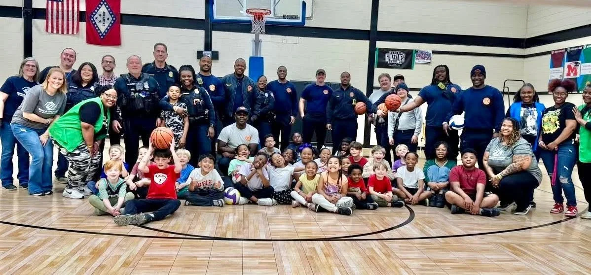 Group photo of children and adults in a gymnasium, some holding basketballs, with banners and flags hanging on the walls.