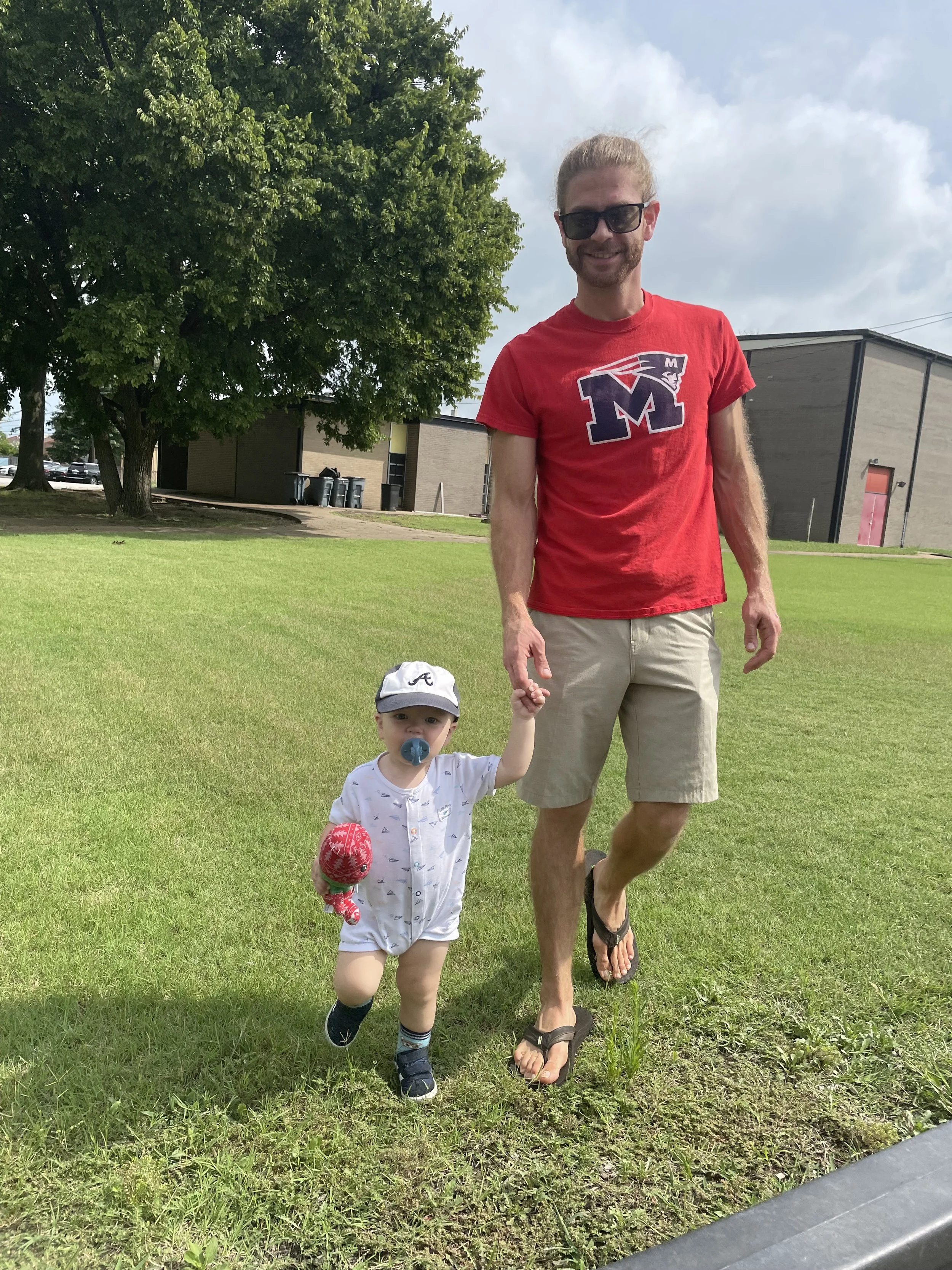 A man with sunglasses in a red Memphis t-shirt holding the hand of a young child who has a pacifier and is wearing a white shirt, shorts, a gray cap, and socks with shoes. They are outdoors on a grassy area with trees and buildings in the background,