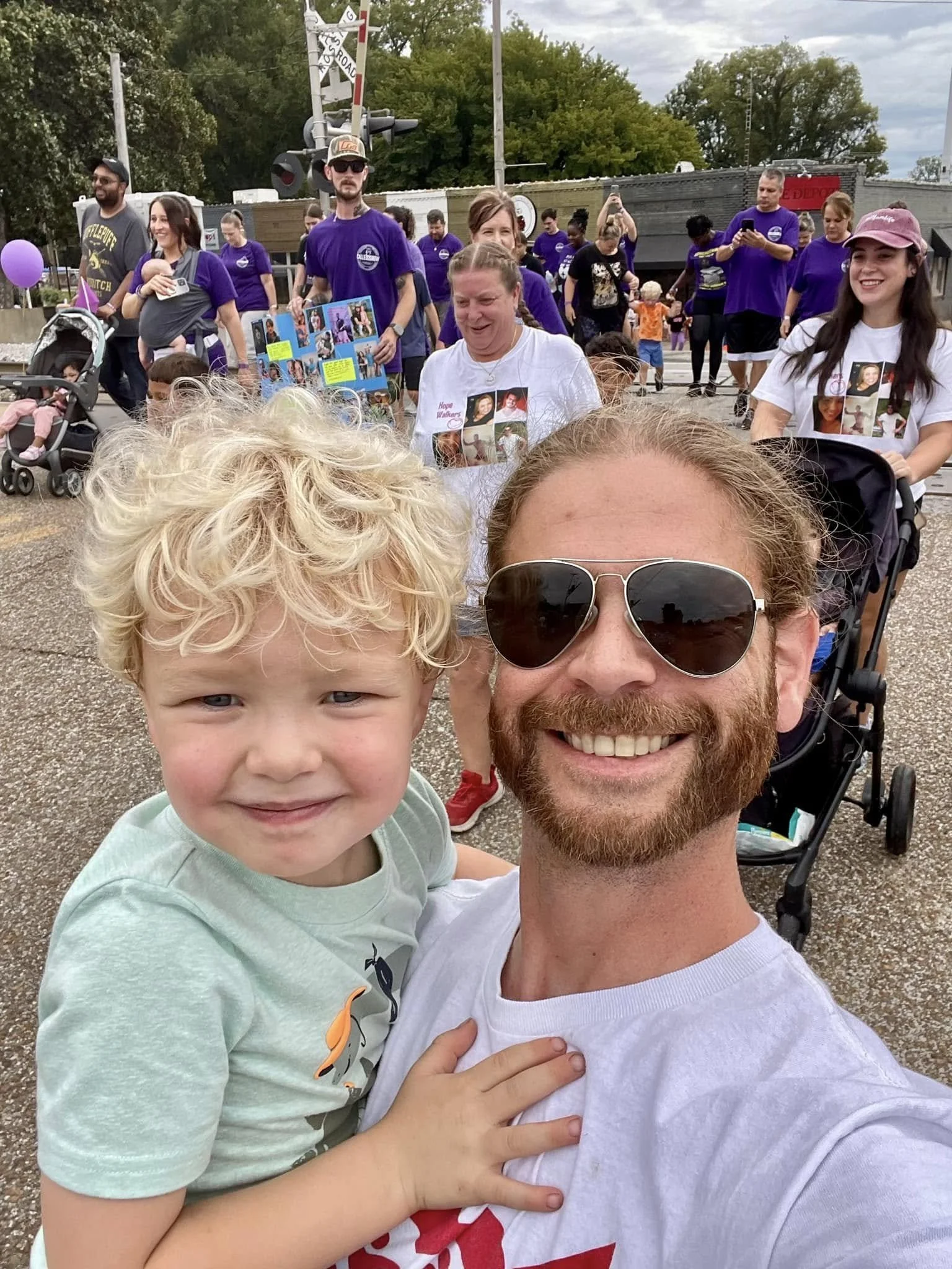 A man with a beard and sunglasses taking a selfie with a young blonde boy with curly hair at an outdoor event. In the background, there are people walking and gathering, some wearing purple shirts, and a train with railroad crossing signals.