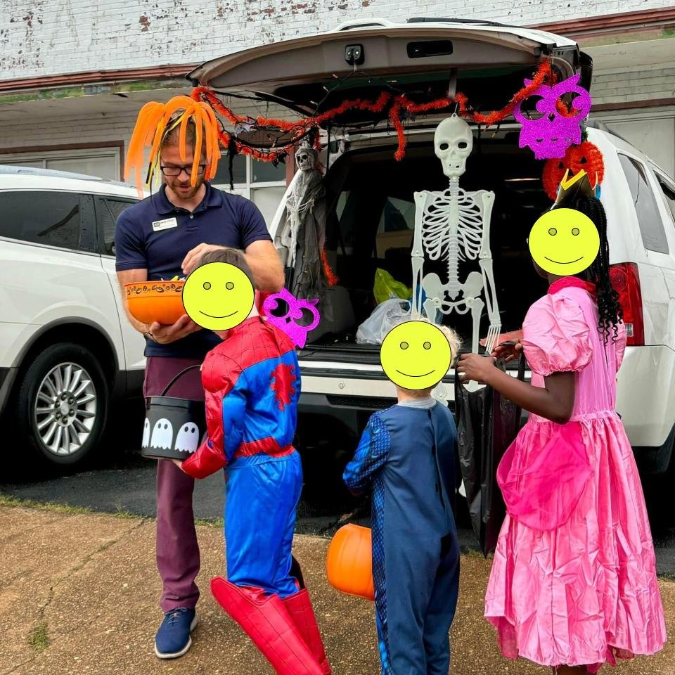A man and three children dressed in Halloween costumes collecting candies from a decorated vehicle trunk. The man is wearing a wig with orange dreadlocks, and the children are dressed as Spider-Man and princesses. The vehicle trunk is decorated with 