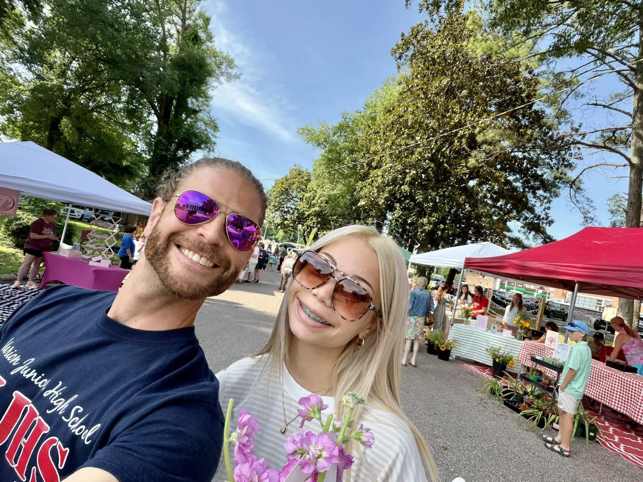 A man with pink sunglasses and a woman with dark sunglasses smiling, at an outdoor market with tents and trees in the background.