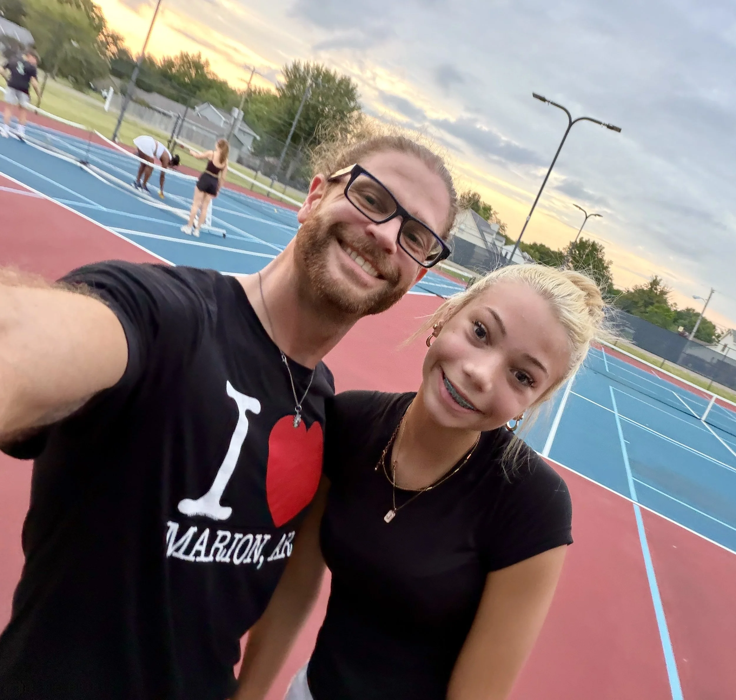 Close-up selfie of two smiling people on a colorful outdoor tennis court during sunset, with other players warming up in the background.