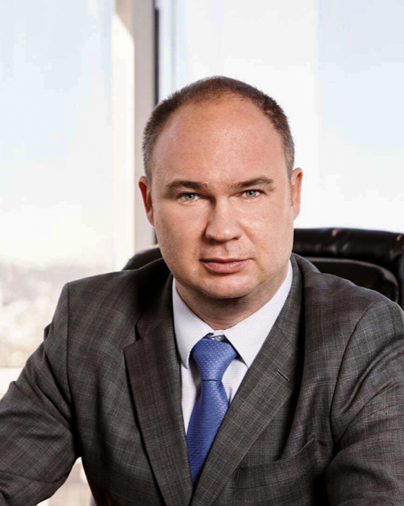 A man in a dark gray suit and blue tie sitting at a desk in an office, with a window in the background.